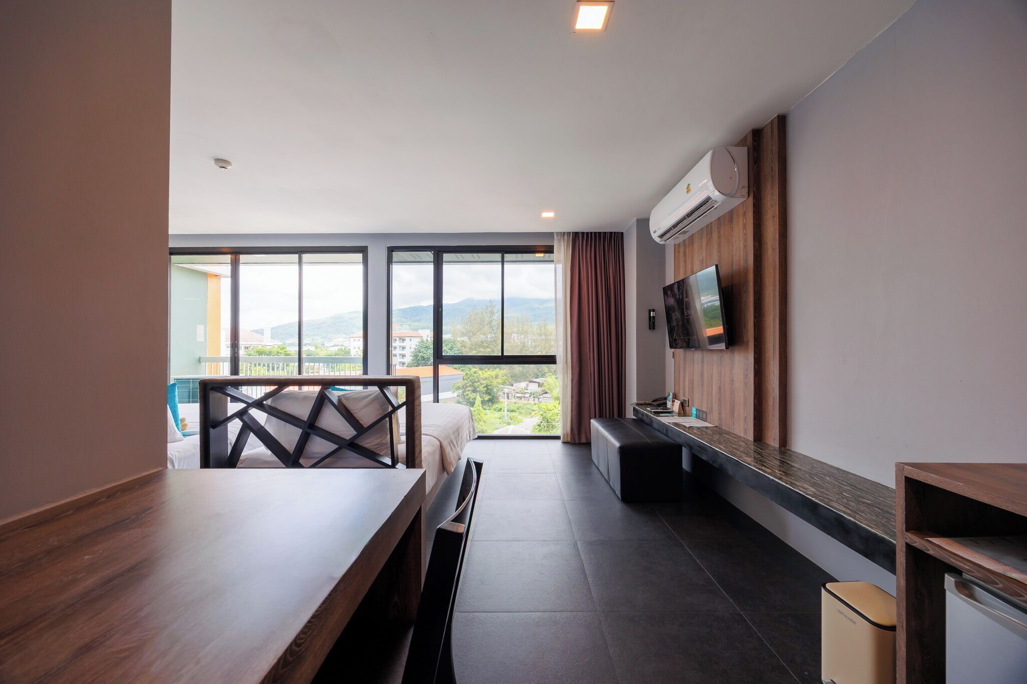A modern hotel room with a large window, a flat-screen TV, a wooden dining table, and a built-in bench.