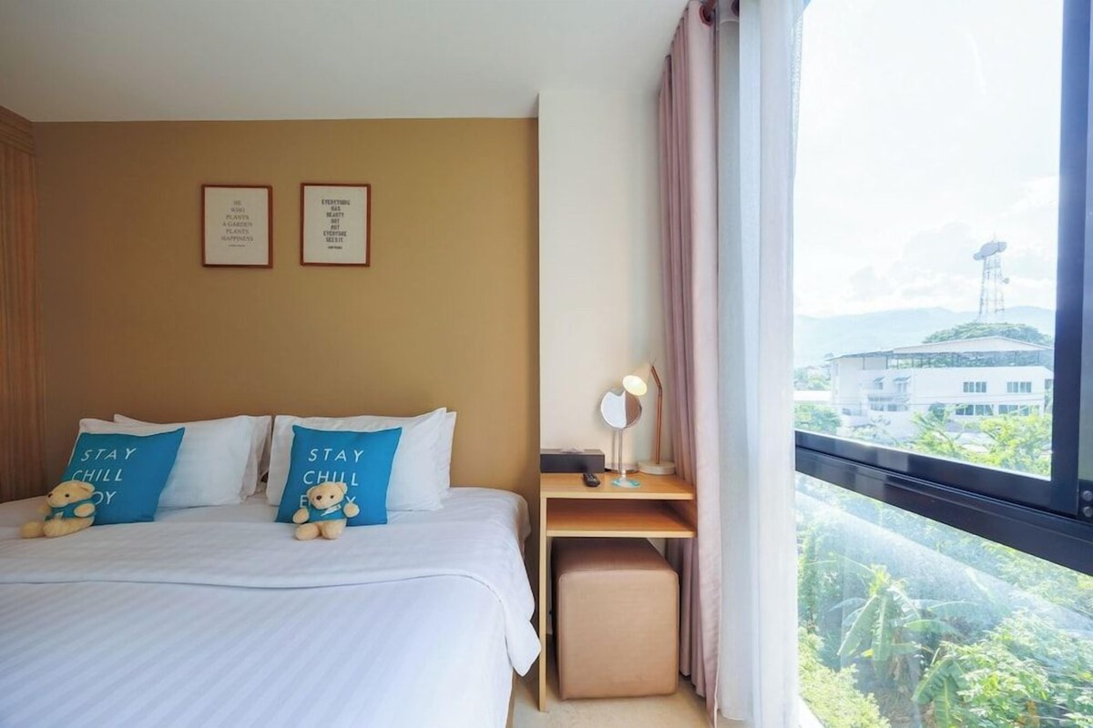 Habitación de hotel con cama, almohadas, mesita de noche, lámpara y ventana con vistas a edificios y zonas verdes.
