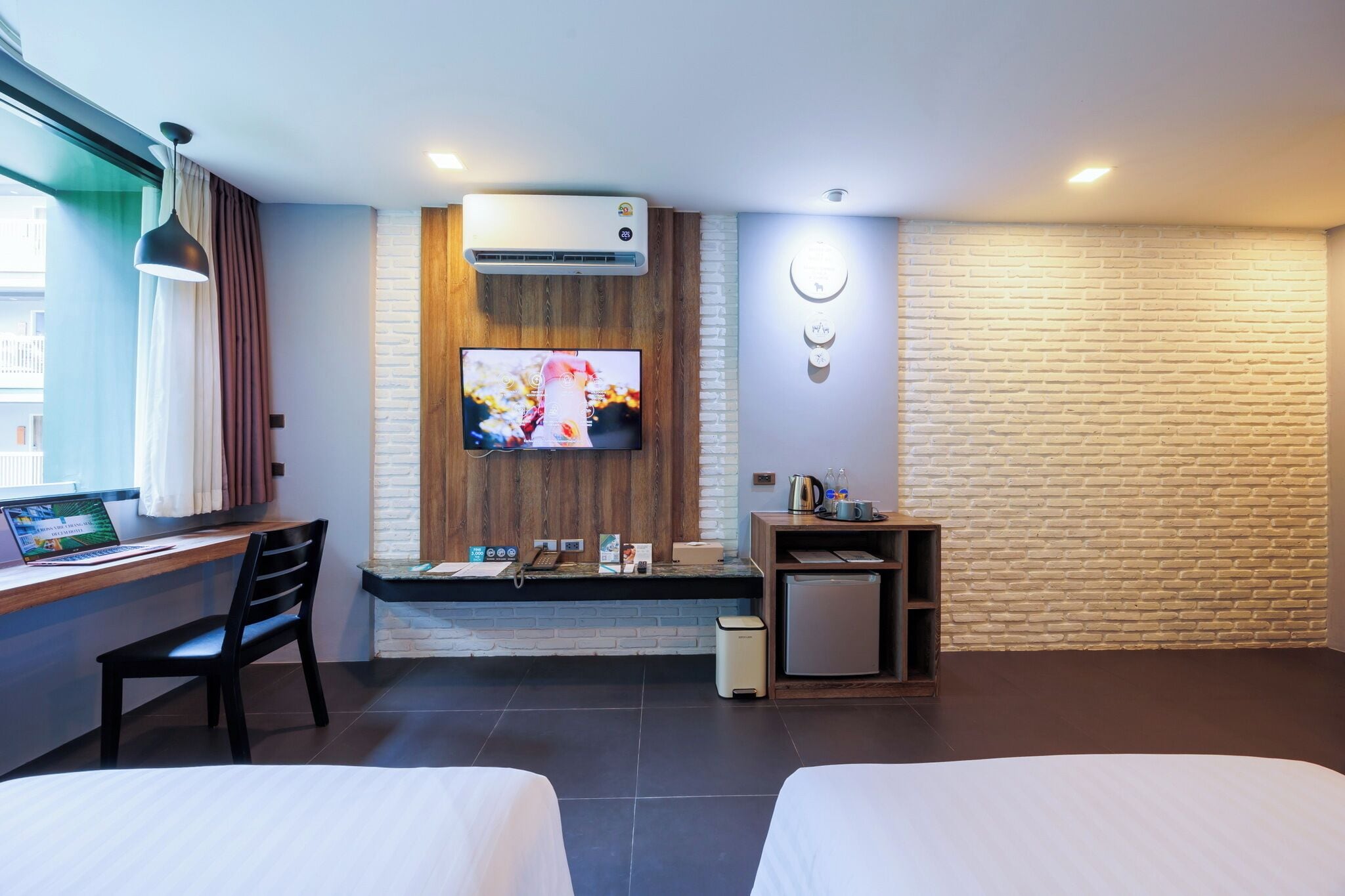 A hotel room with a bed, a desk with a computer, a TV, a small fridge, and a brick-patterned wall.