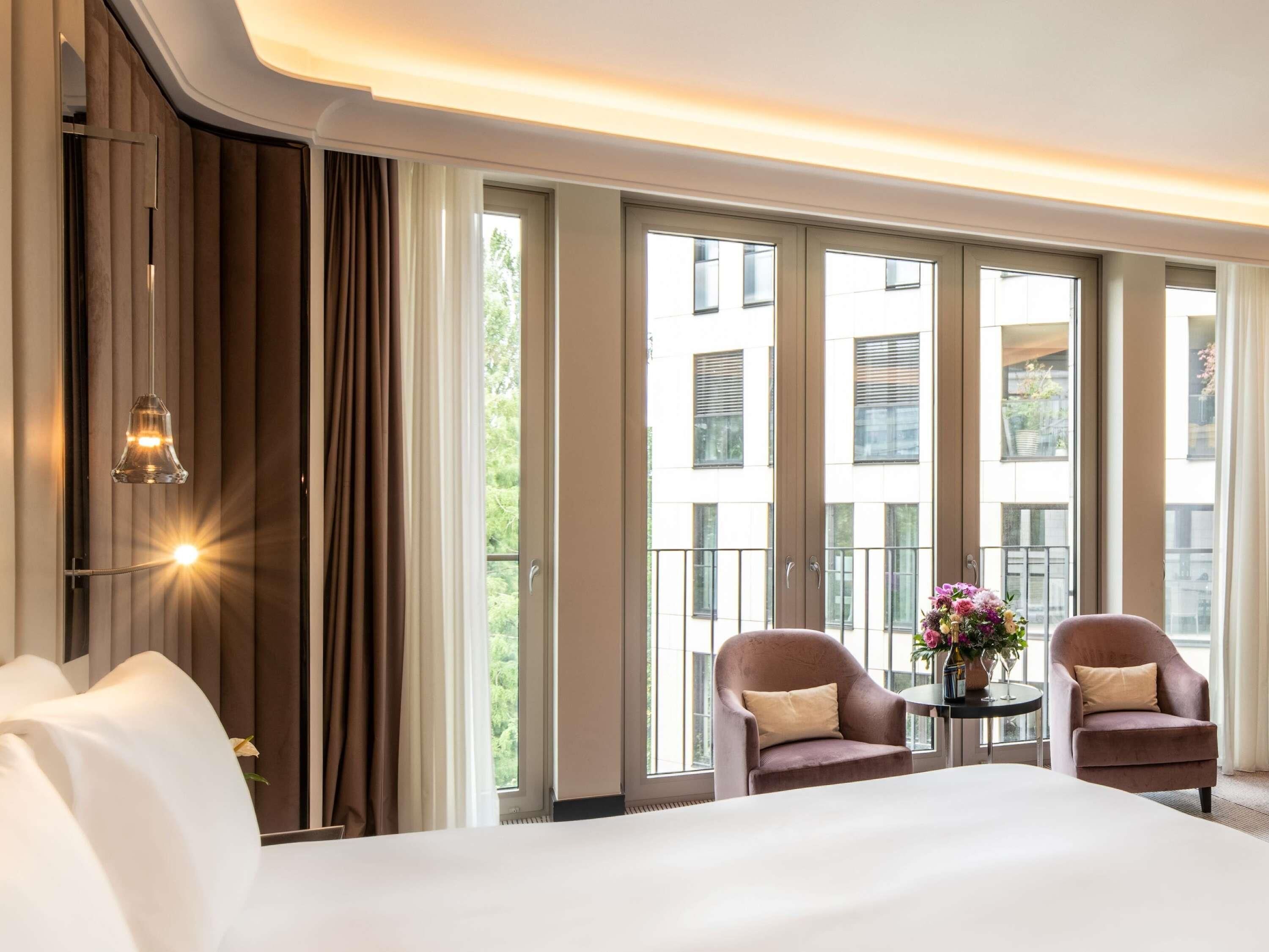 A hotel room with a large bed, two armchairs, a small table with a vase of flowers, and large windows offering a view of the city.