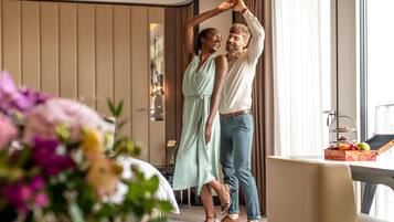 A couple dancing in a hotel room with a bed, a table with fruit, and a view of the outdoors.