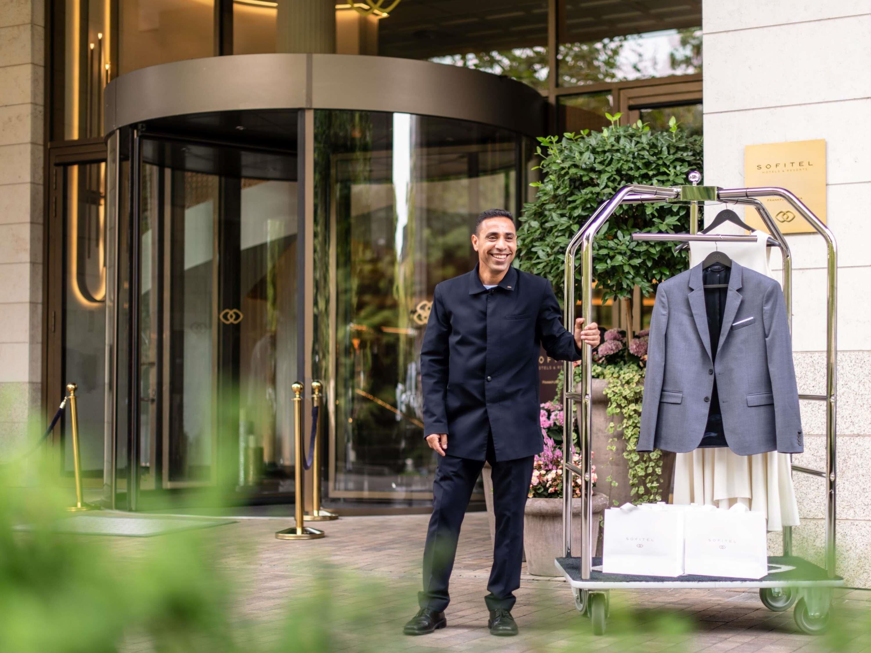 Un hombre con traje de pie junto a una puerta giratoria, con un perchero que muestra un traje y bolsas de compras.