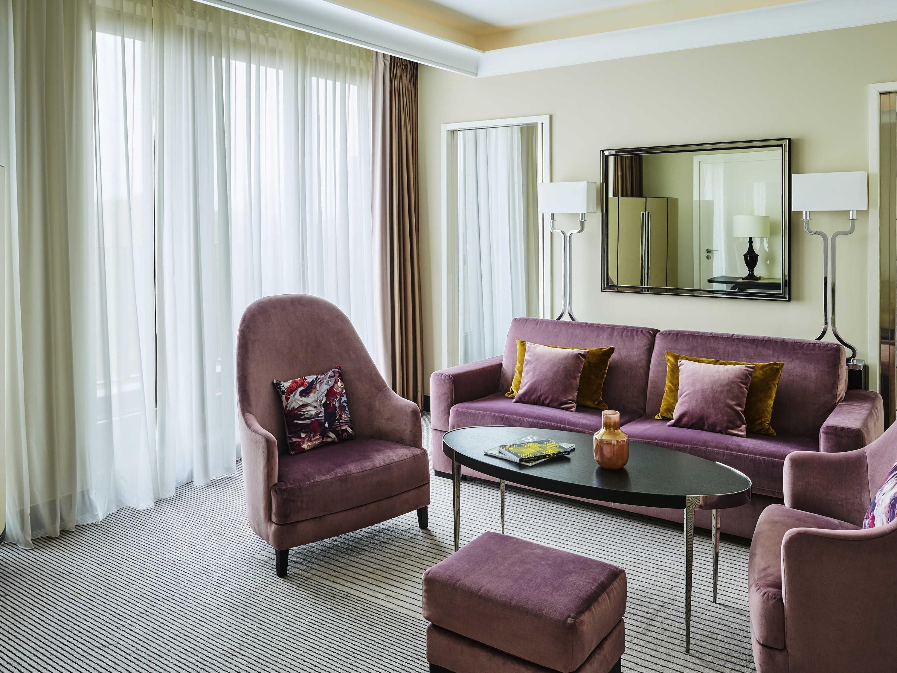 A modern hotel room with a purple sofa, a matching armchair, a small ottoman, and a coffee table. There is a large mirror on the wall and two floor lamps.