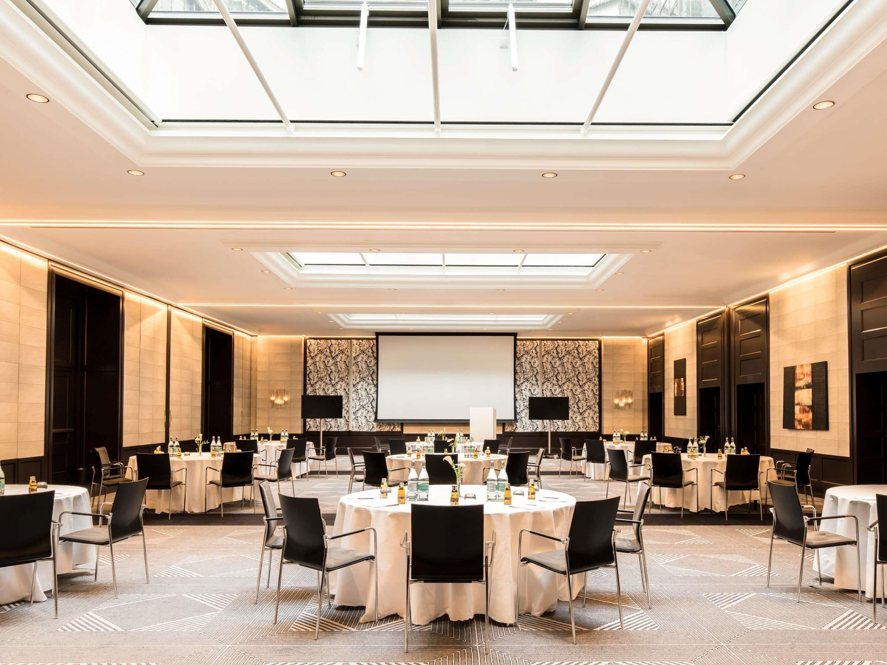 A spacious conference room with round tables, black chairs, and a large screen at the front.