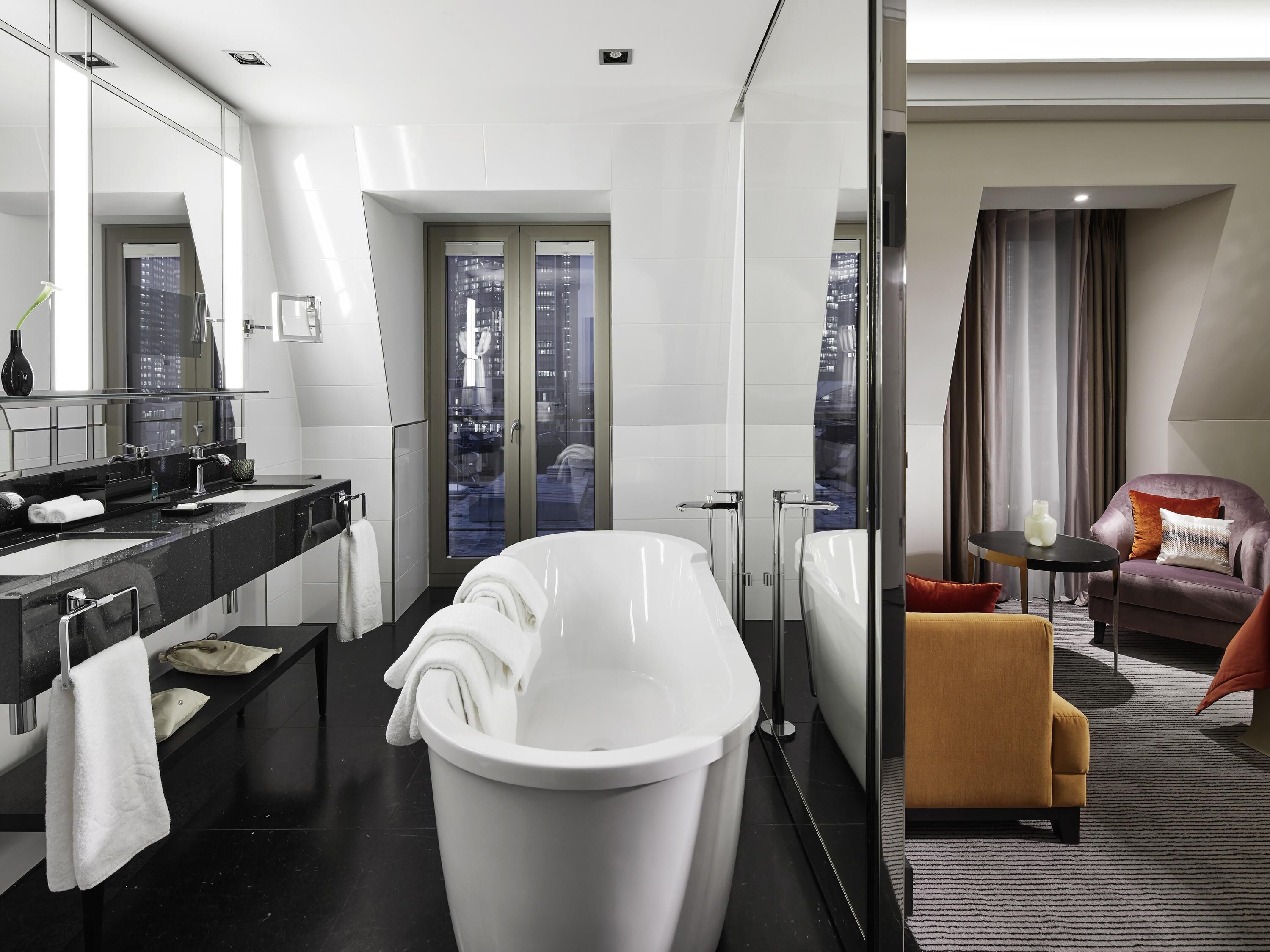 Un bagno moderno con una vasca, uno specchio grande e vista sulla città attraverso una porta in vetro.