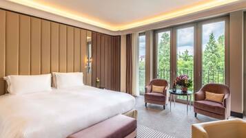 A hotel room with a large bed, a small table with chairs, and a balcony with a view of trees.
