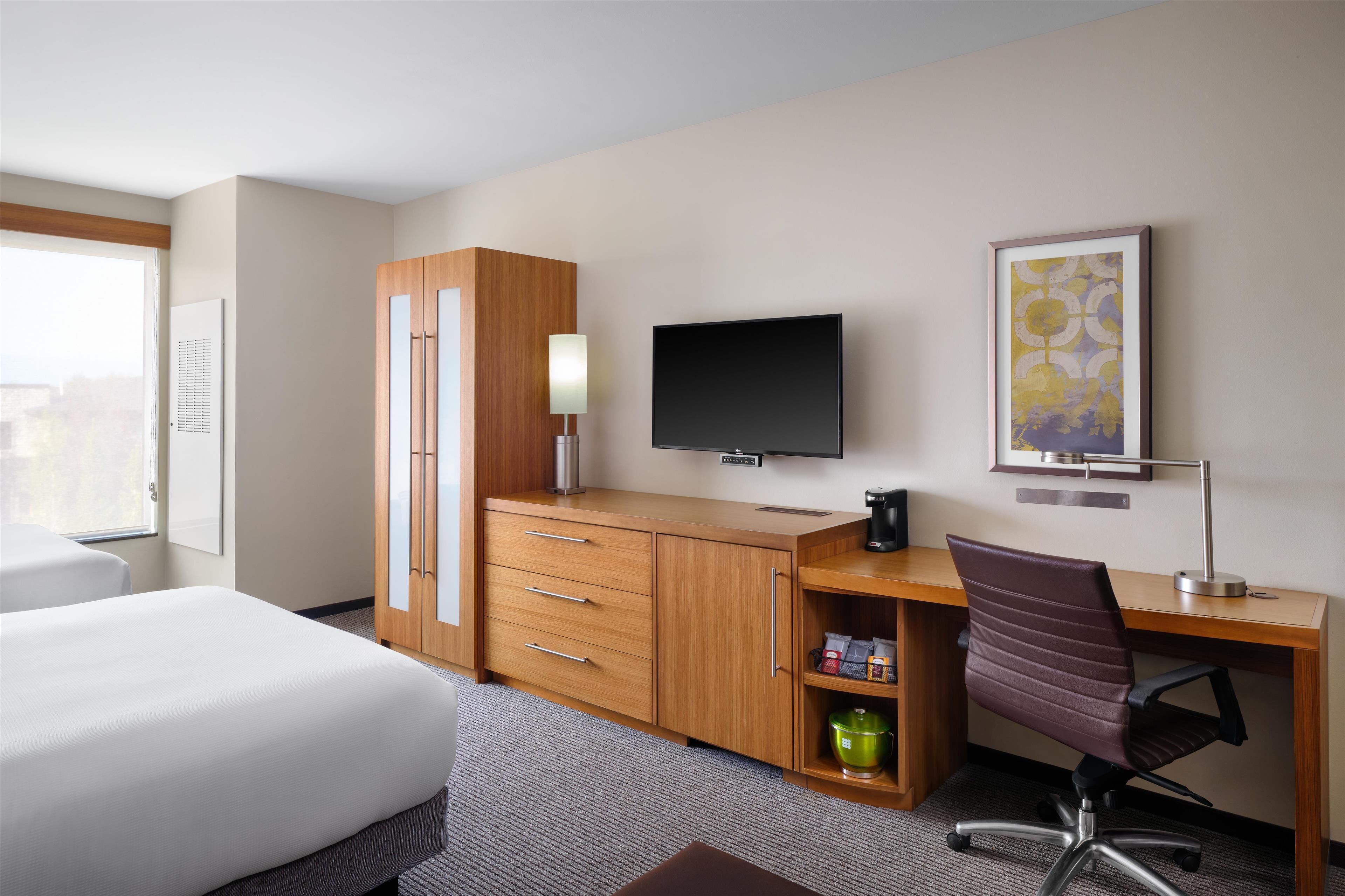 A hotel room with a bed, a wooden desk, a television, a chair, and a shelf with toiletries.