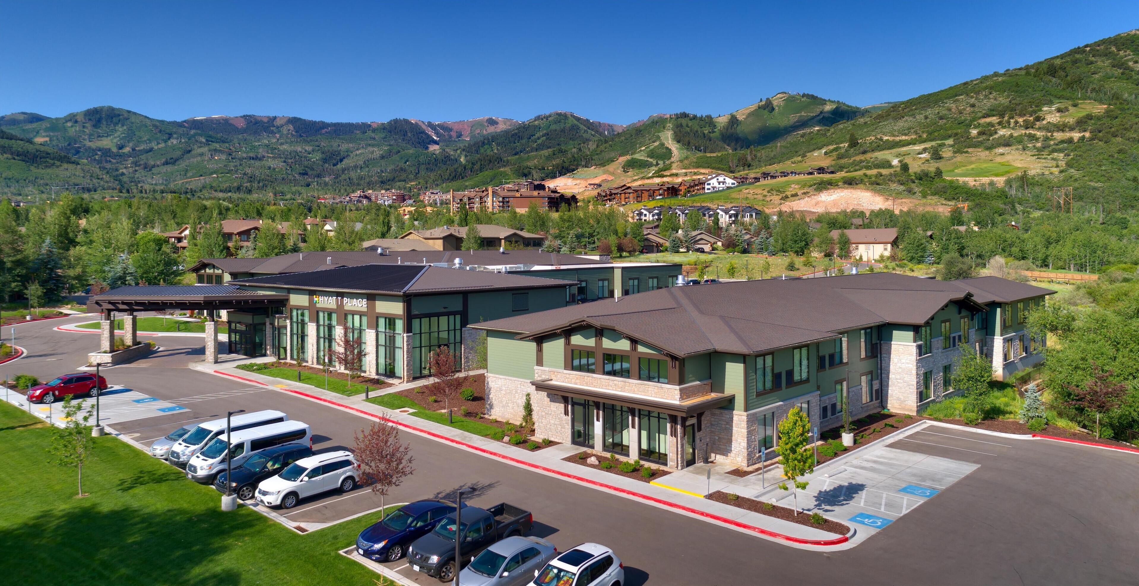 A modern hotel with a parking lot, surrounded by mountains and greenery.
