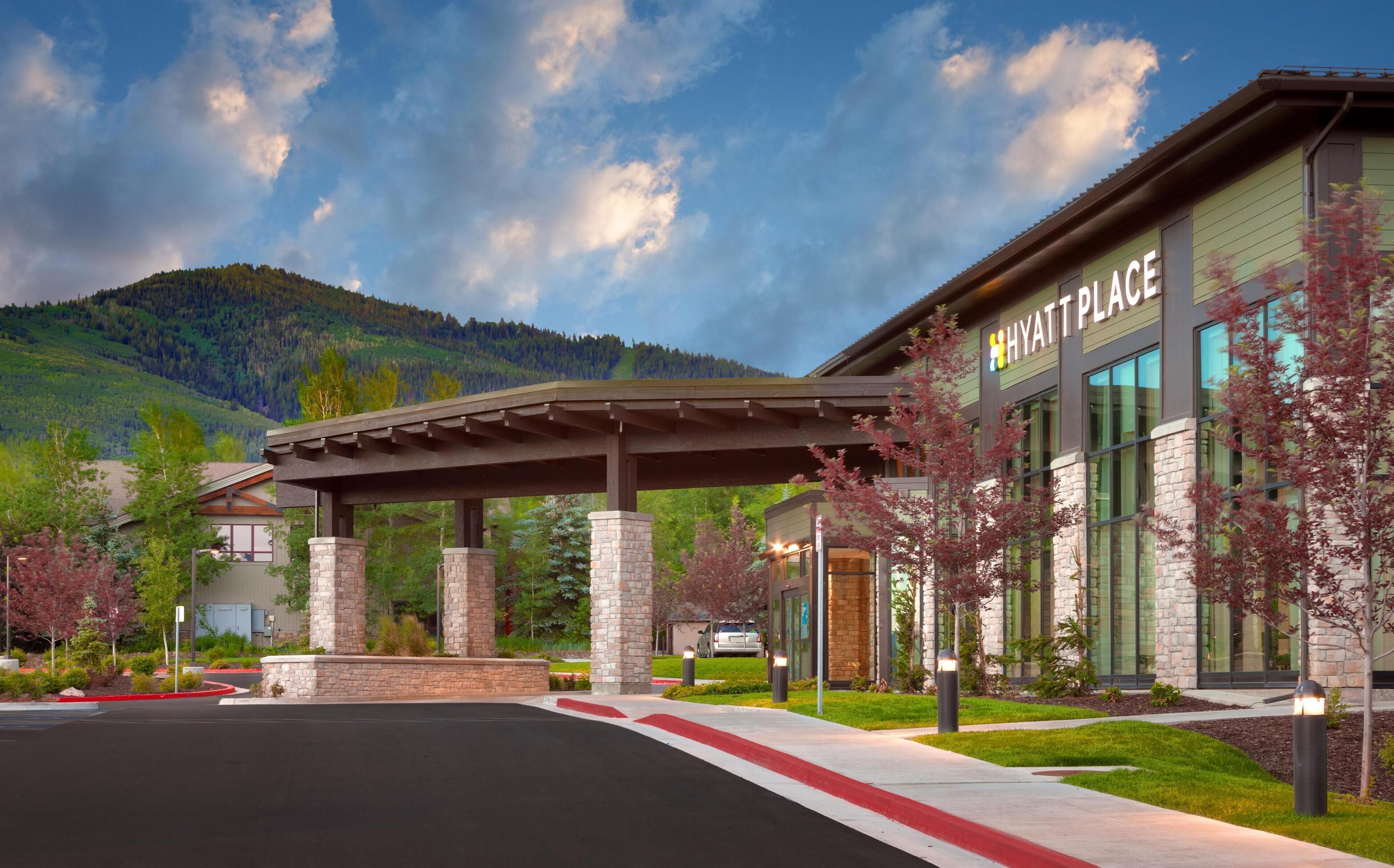 A Hyatt Place building with a covered entrance, stone pillars, and a view of a mountainous landscape.