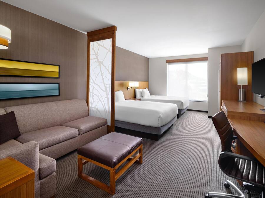 A hotel room with a sofa, two beds, a desk, a TV, and a large window.