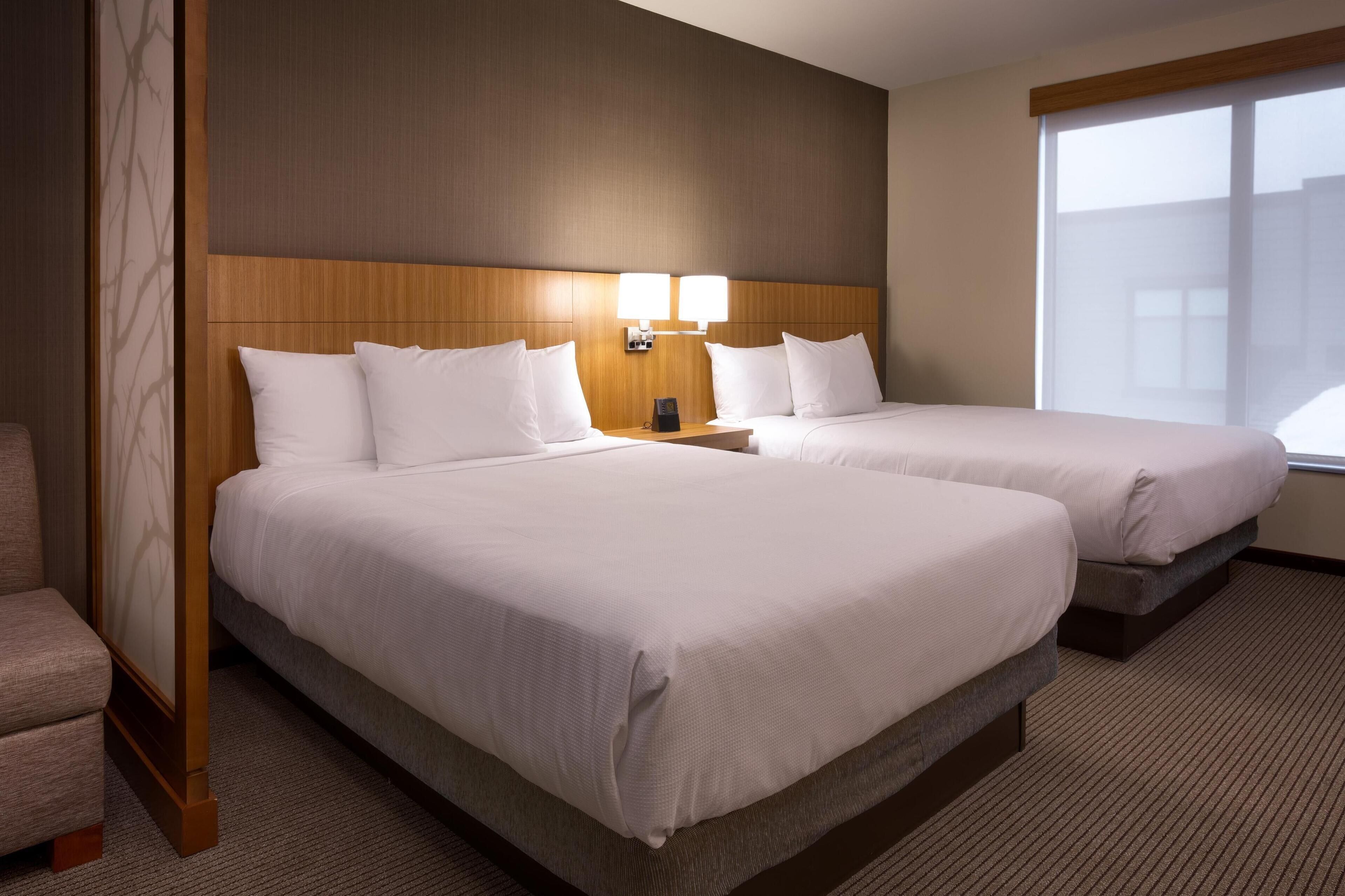 A hotel room with two beds, a wooden headboard, a nightstand with a lamp, and a large window.
