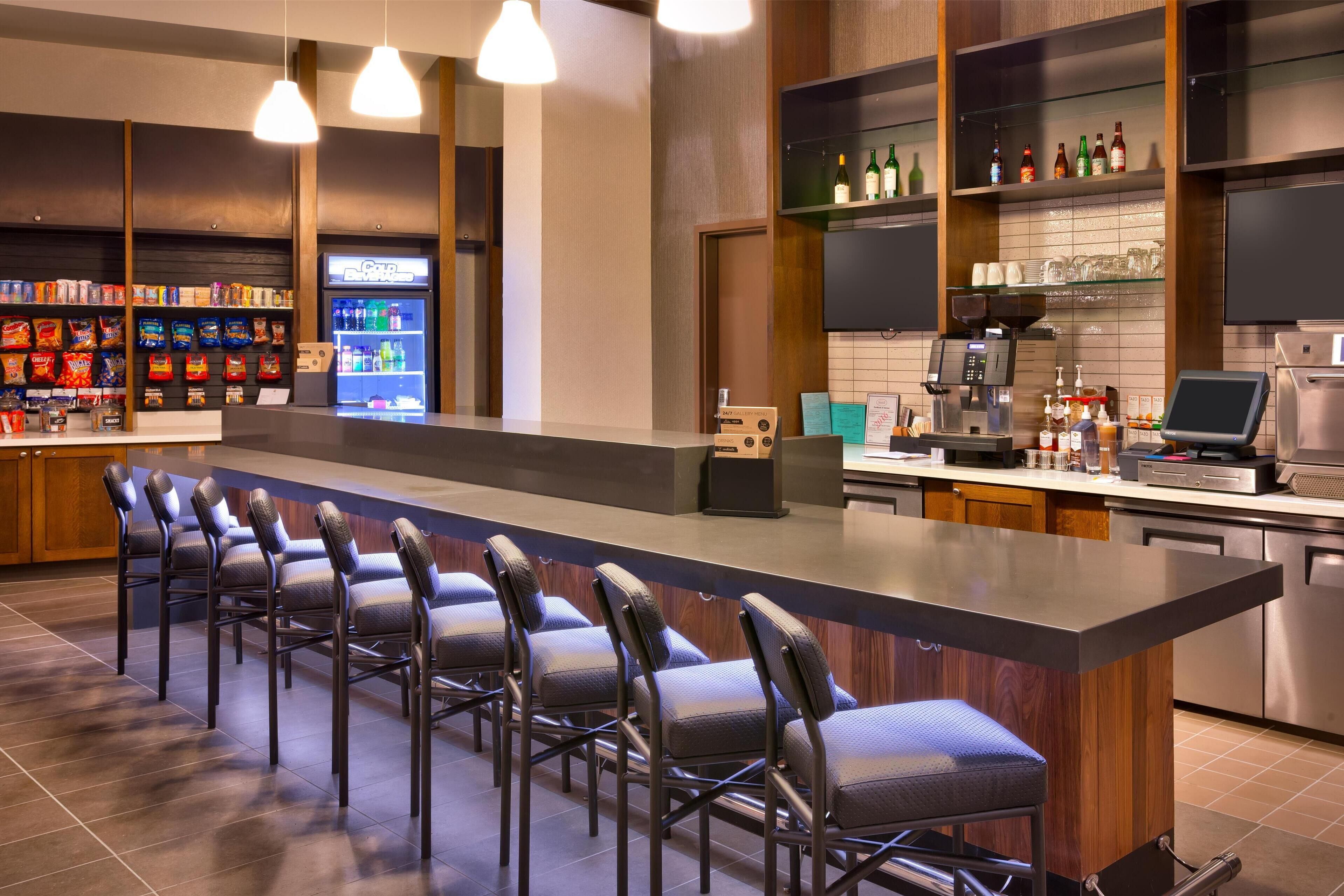 A modern bar area with a long counter, multiple bar stools, a beverage dispenser, and a coffee machine.