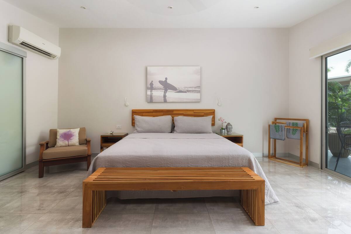 A modern bedroom with a large bed, wooden headboard, and a bench. There is a nightstand with a lamp and a small table with a plant. A large window provides a view of the outdoors.