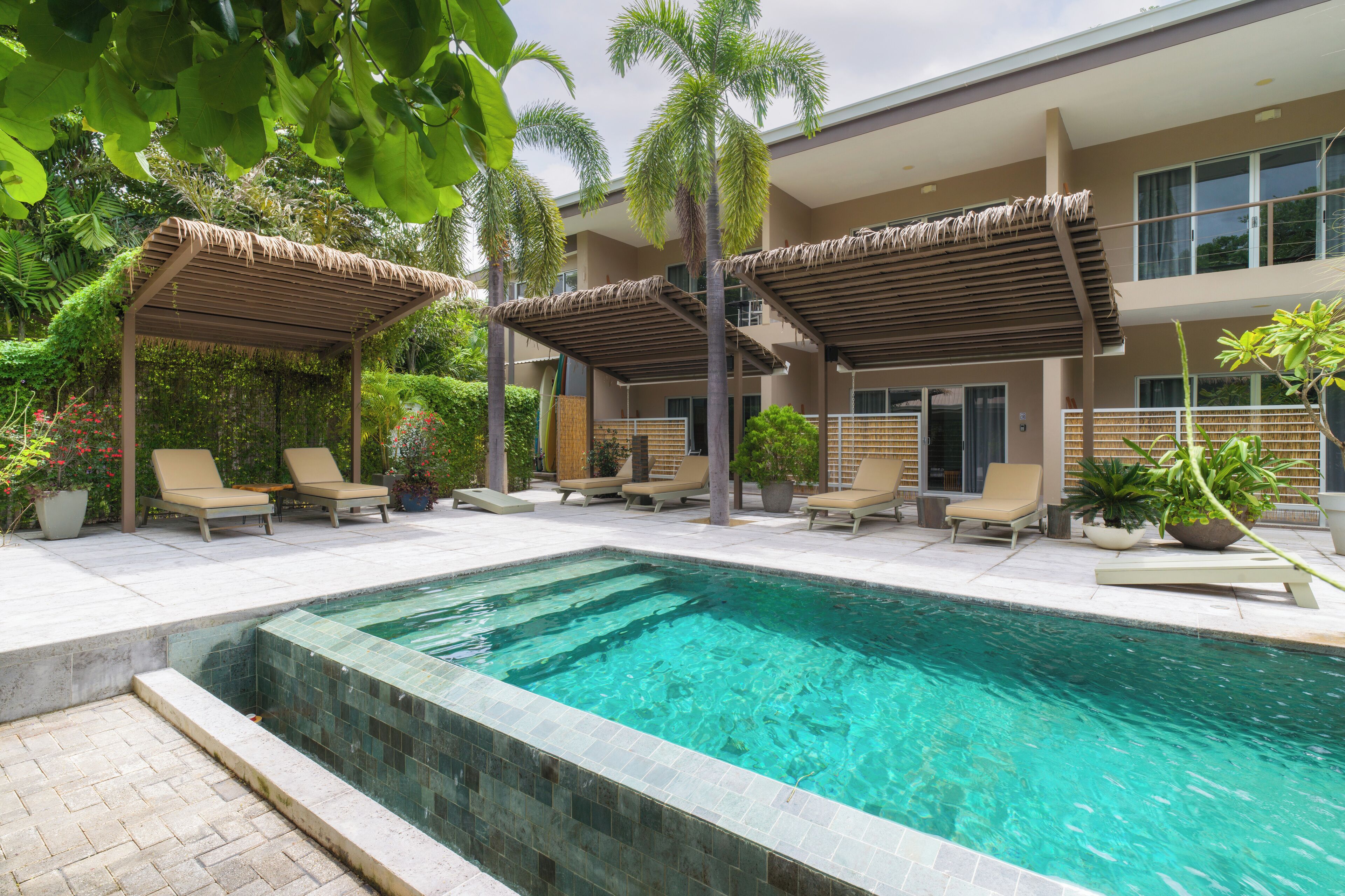 A swimming pool with lounge chairs and a covered seating area, surrounded by greenery and a building with multiple balconies.
