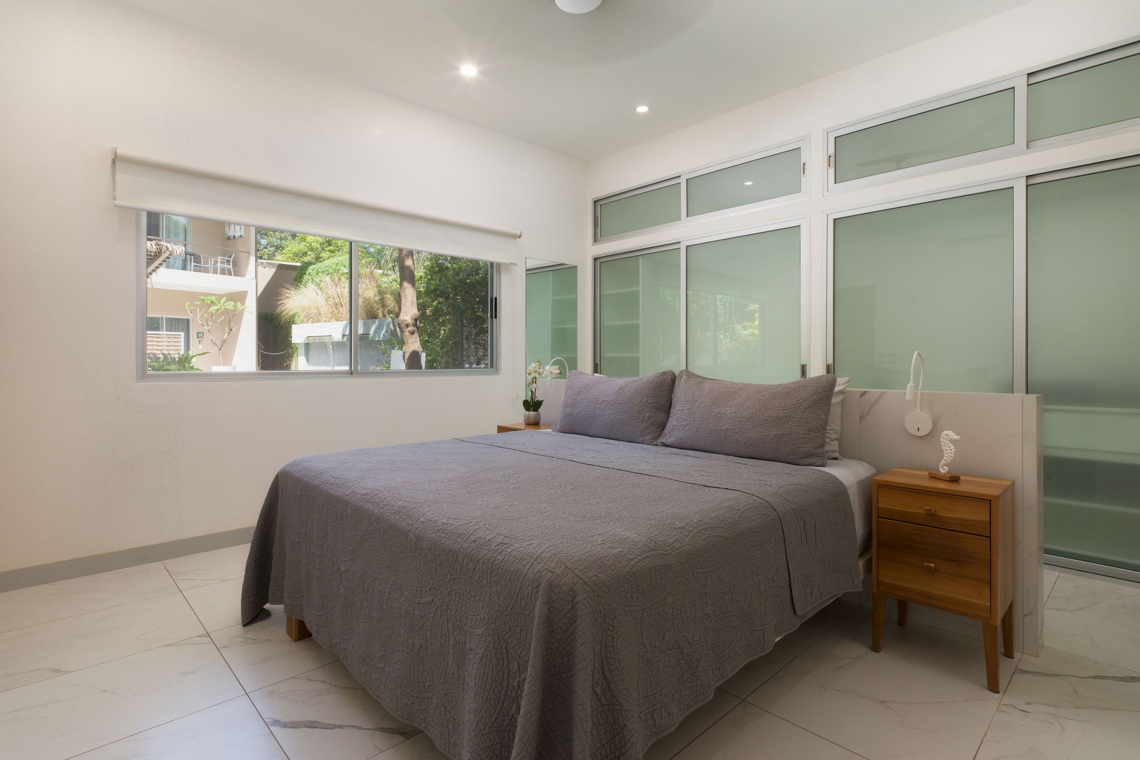 Un dormitorio con una cama grande, una mesita de noche, una ventana y vistas al verdor exterior.