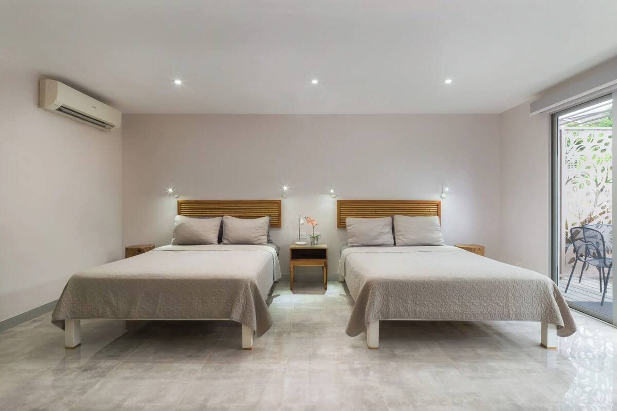 A modern hotel room with two beds, a small table with a lamp, and a large window with a view of greenery.