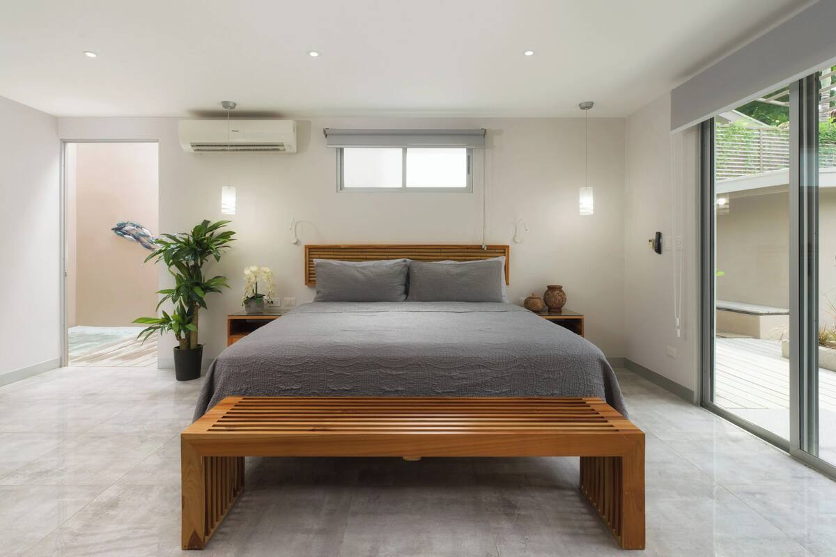 A modern bedroom with a wooden bed, a bench, a potted plant, and a view through large sliding doors.