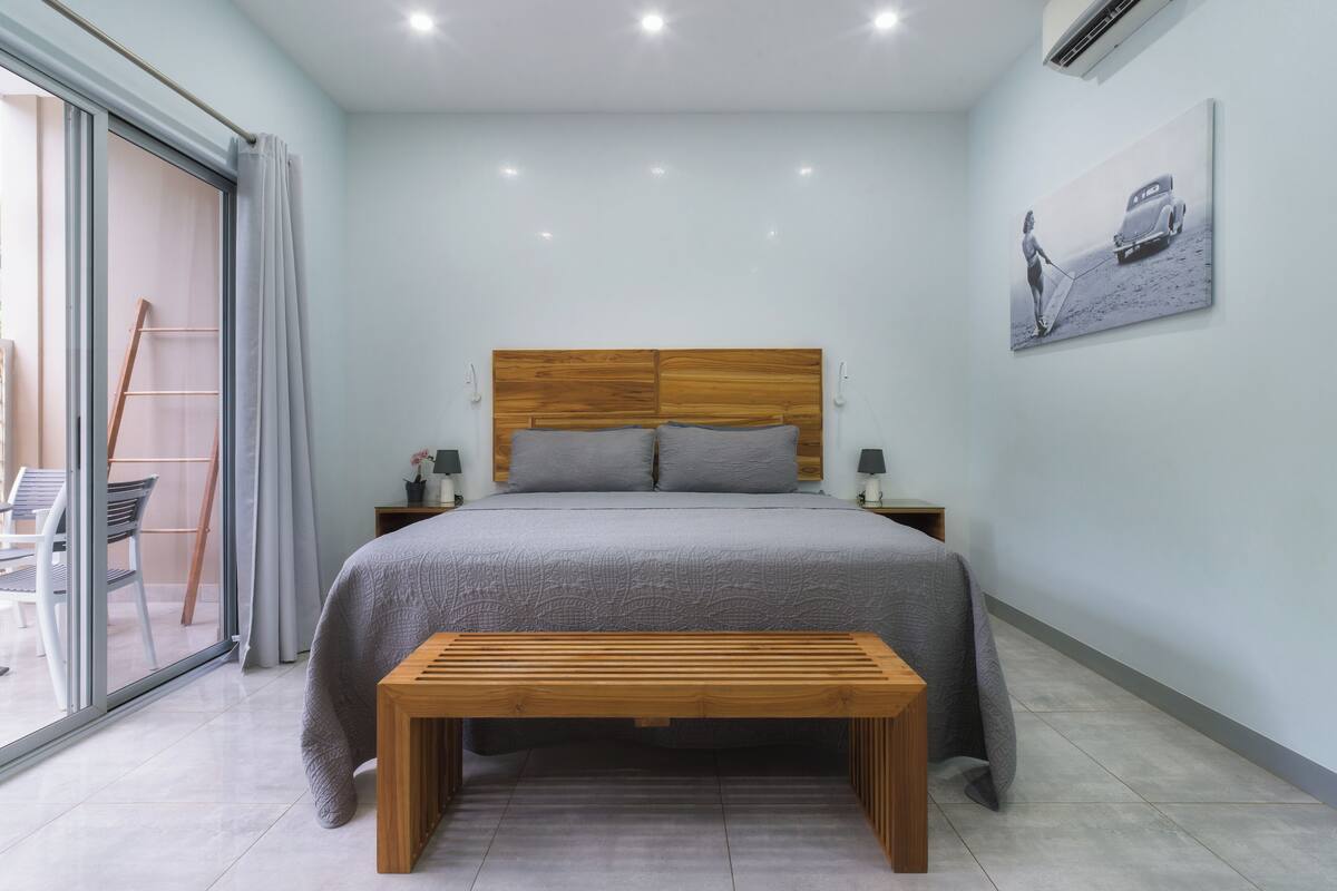A bedroom with a bed, wooden headboard, grey bedding, a bench, a nightstand, a lamp, and a painting on the wall.