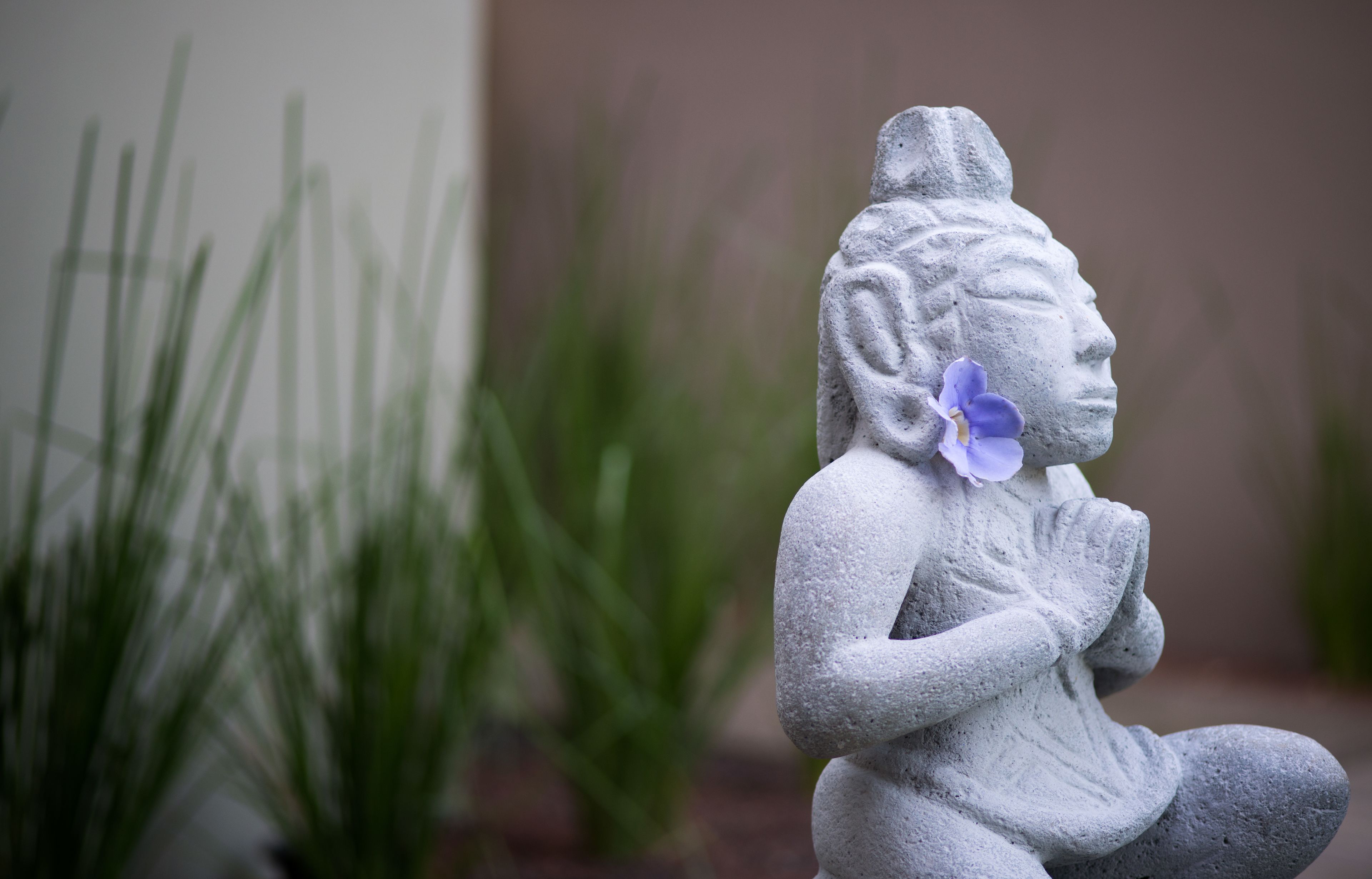 A stone statue of a seated figure with a flower in its hair, set against a backdrop of green plants.