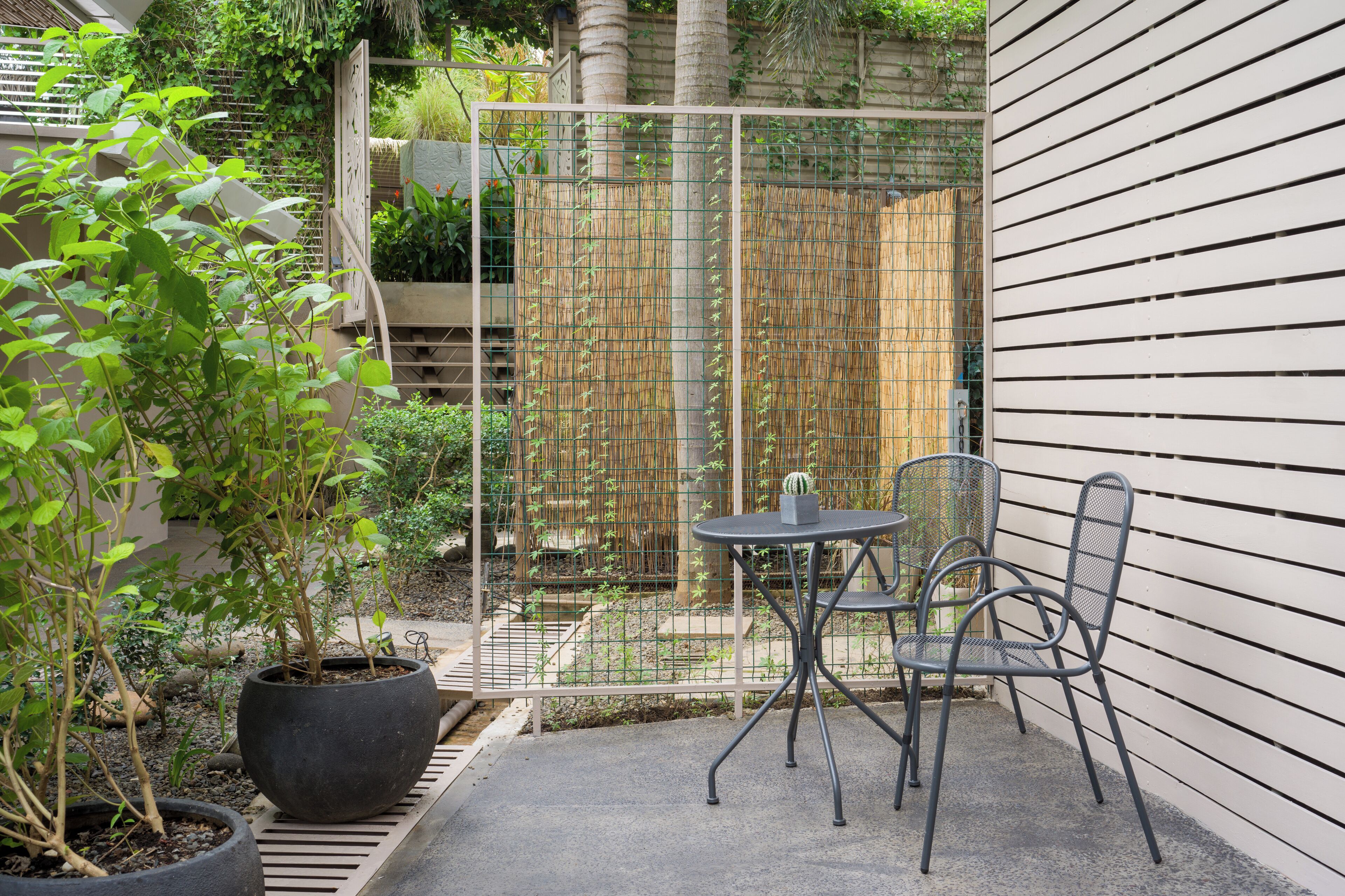 A small outdoor patio area with a metal table and chairs, surrounded by potted plants and a bamboo fence.