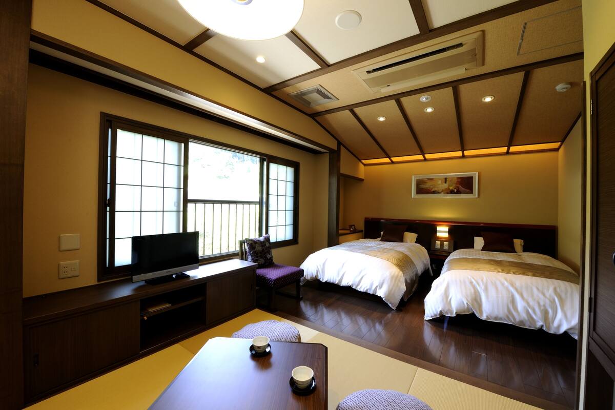 A hotel room with a traditional Japanese aesthetic, featuring tatami flooring, a low table with a tea set, a television, and two beds with headboards.