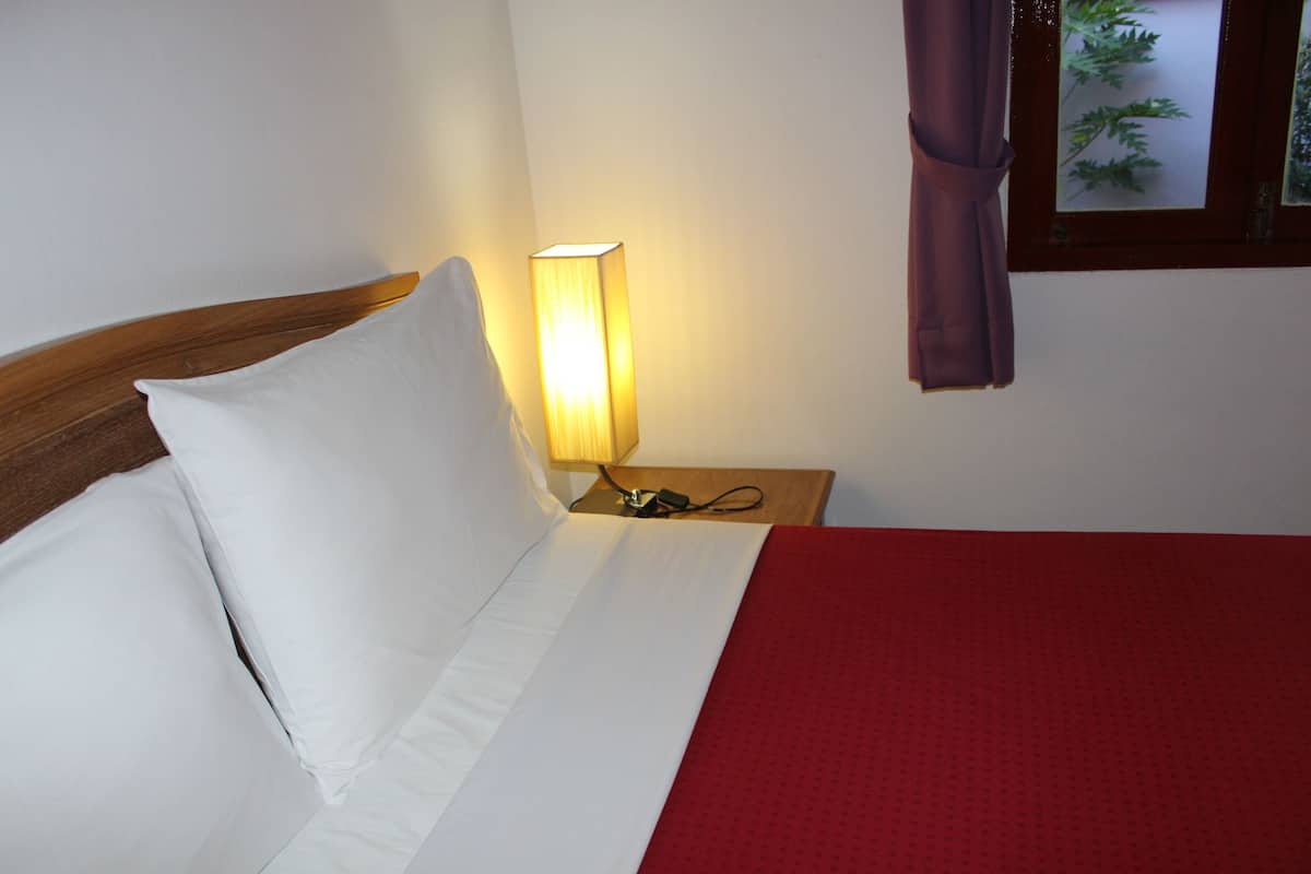 A neatly made bed with white linens, a red blanket, and a wooden headboard. There is a bedside table with a lamp and a power outlet.