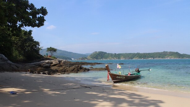 A boat named 'The Bavarian' on a sandy beach with clear water, rocky outcrops, and a forested hill in the background. - The Happy Elephant Resort (Rawai)