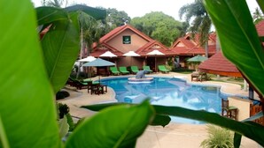 A pool area with lounge chairs and umbrellas, surrounded by greenery and a building with a red roof. - The Happy Elephant Resort (Rawai)