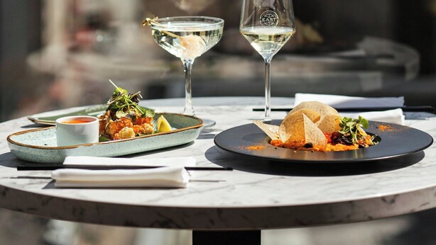 A dining table with plates of food and two glasses of white wine, set against a cityscape view. - Renaissance Montreal Downtown Hotel (Montreal)