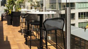 An outdoor terrace with a row of tables and chairs, overlooking a cityscape with modern buildings.