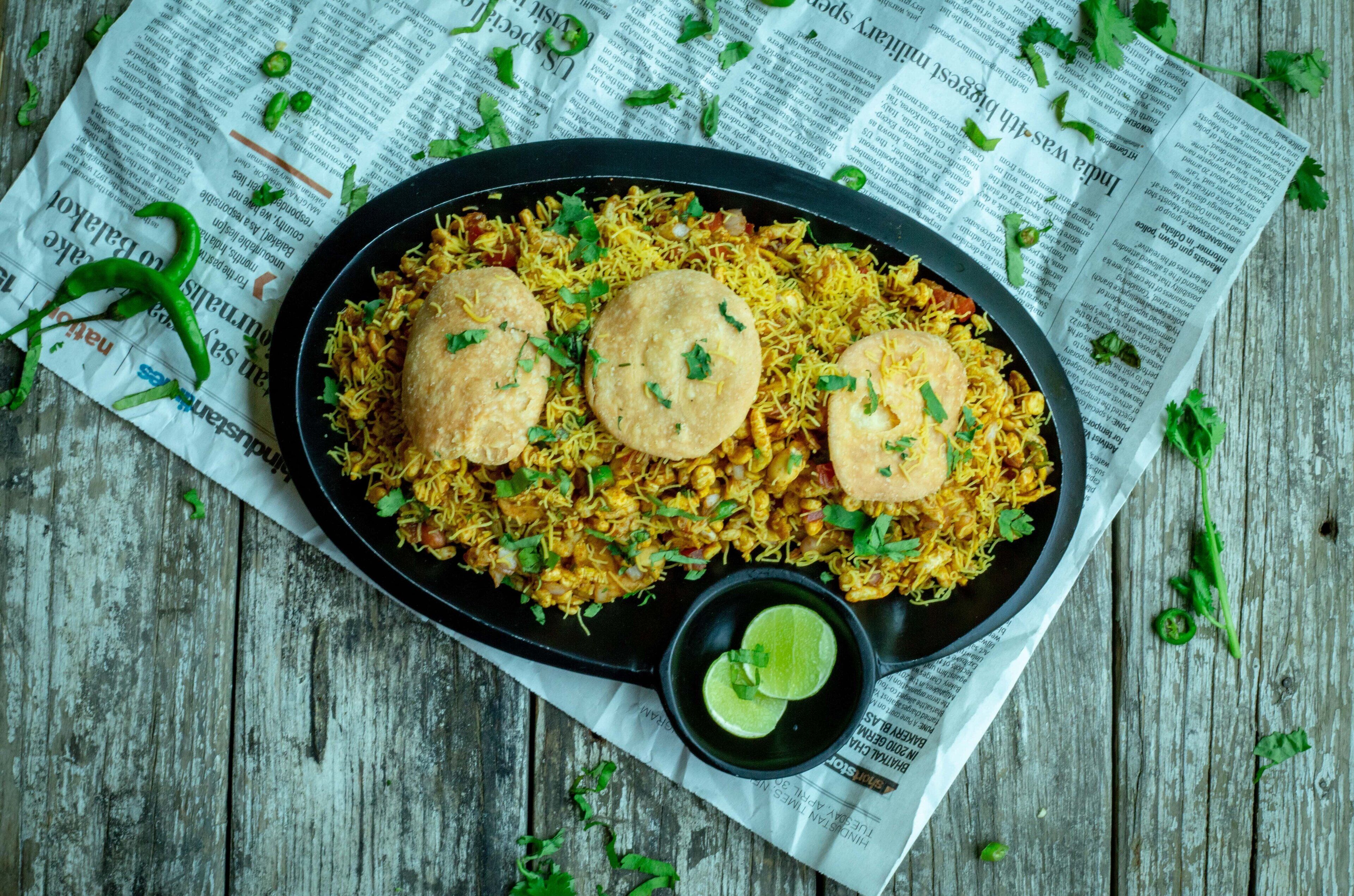 Un plato de biryani con dos panes naan encima, adornado con hierbas, y un recipiente pequeño con cuñas de lima sobre una superficie de madera cubierta con periódico.