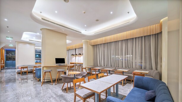 A modern hotel lobby with a mix of seating areas, including sofas and tables, and a large TV screen.