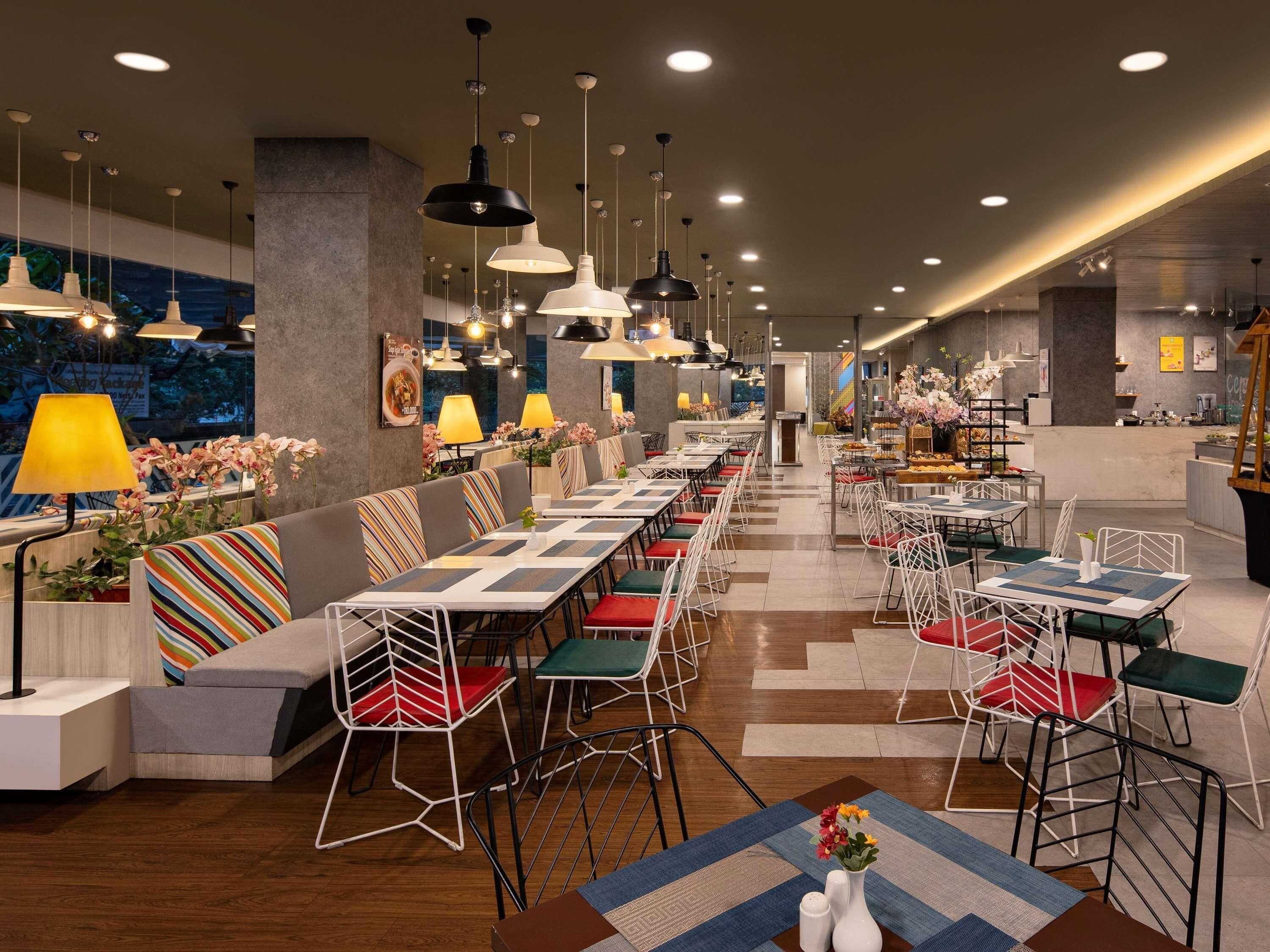 A modern restaurant with long communal tables and individual booths, overhead pendant lighting, and a view of the cityscape through large windows.