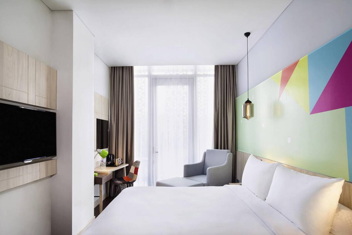A hotel room with a bed, a TV, a desk with a lamp, a chair, and a colorful wall.