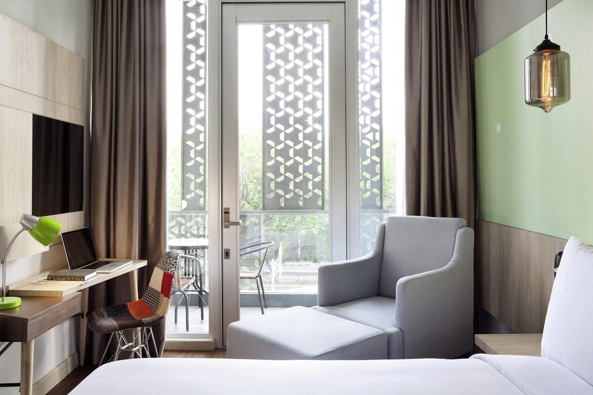 A modern hotel room with a bed, a desk with a laptop, a chair, and a balcony with a view.