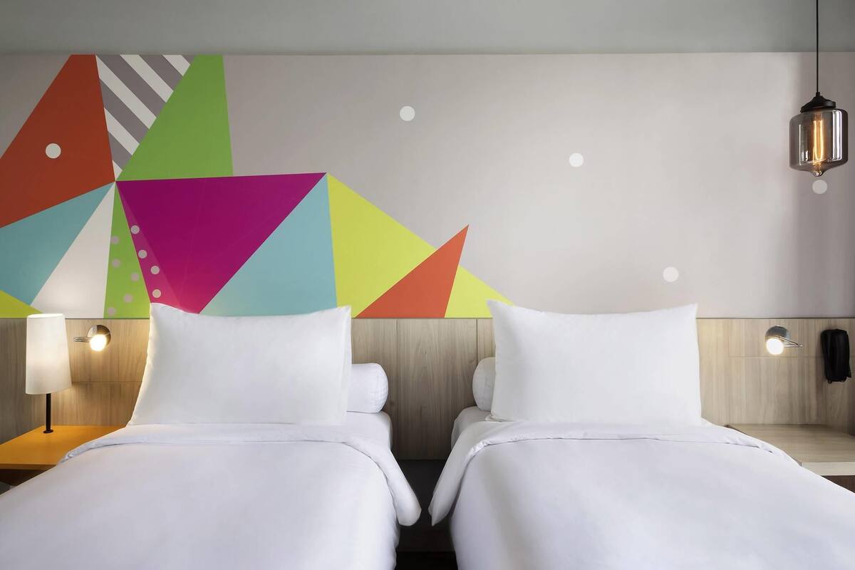 A hotel room with two beds, a colorful geometric wall art, a hanging light fixture, and a bedside table with a lamp.