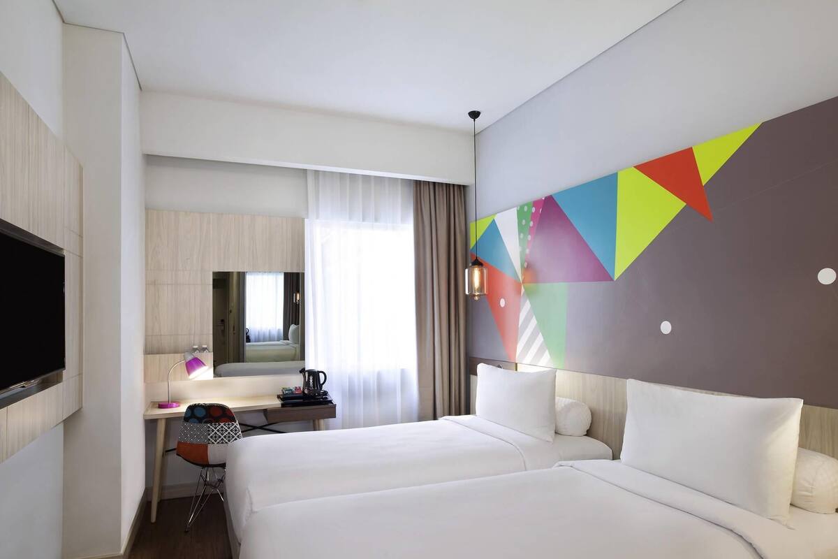 A hotel room with two beds, a desk with a chair, a TV, and a colorful geometric wall design.