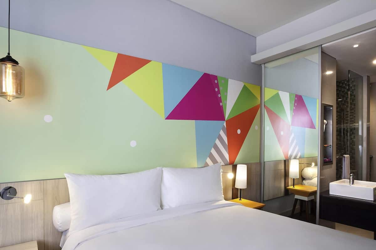 A modern hotel room with a large bed, a bedside table with a lamp, a bathroom with a mirror and sink, and a colorful geometric wall design.