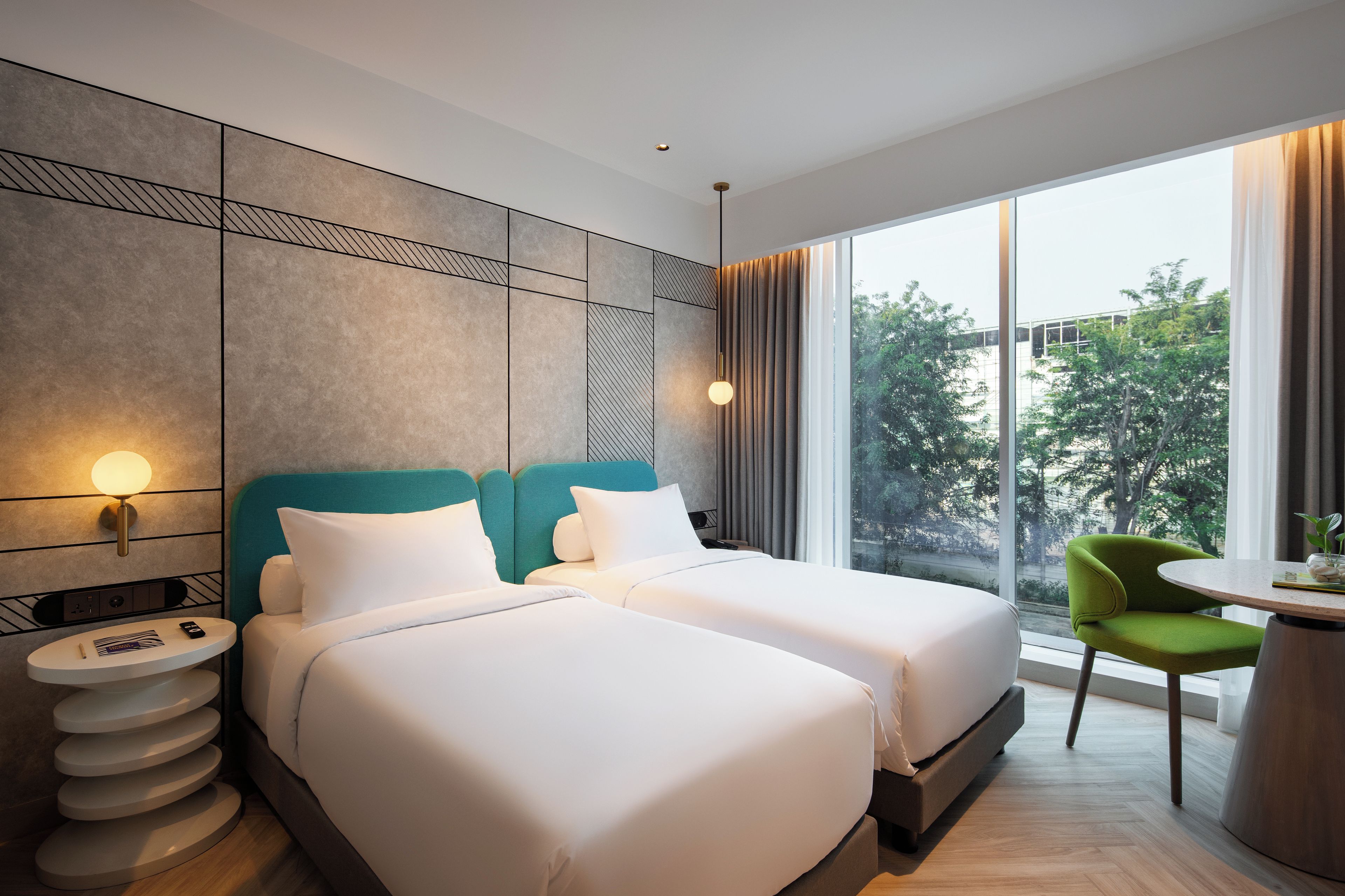 Ein Hotelzimmer mit zwei Betten, einem kleinen Tisch, einem Sessel und einem großen Fenster mit Blick auf Bäume.