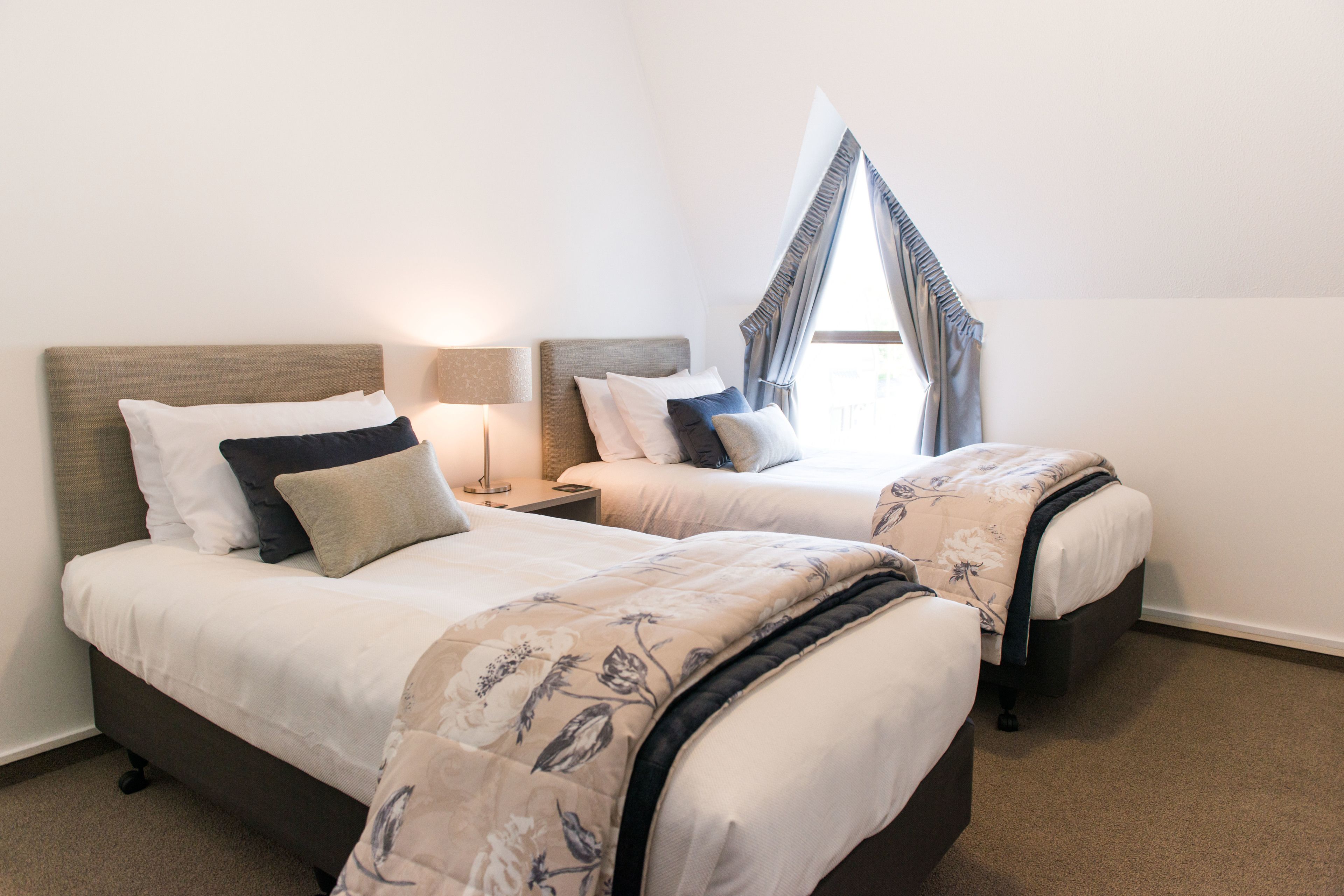 A hotel room with two beds, a nightstand, a lamp, and a window with curtains.