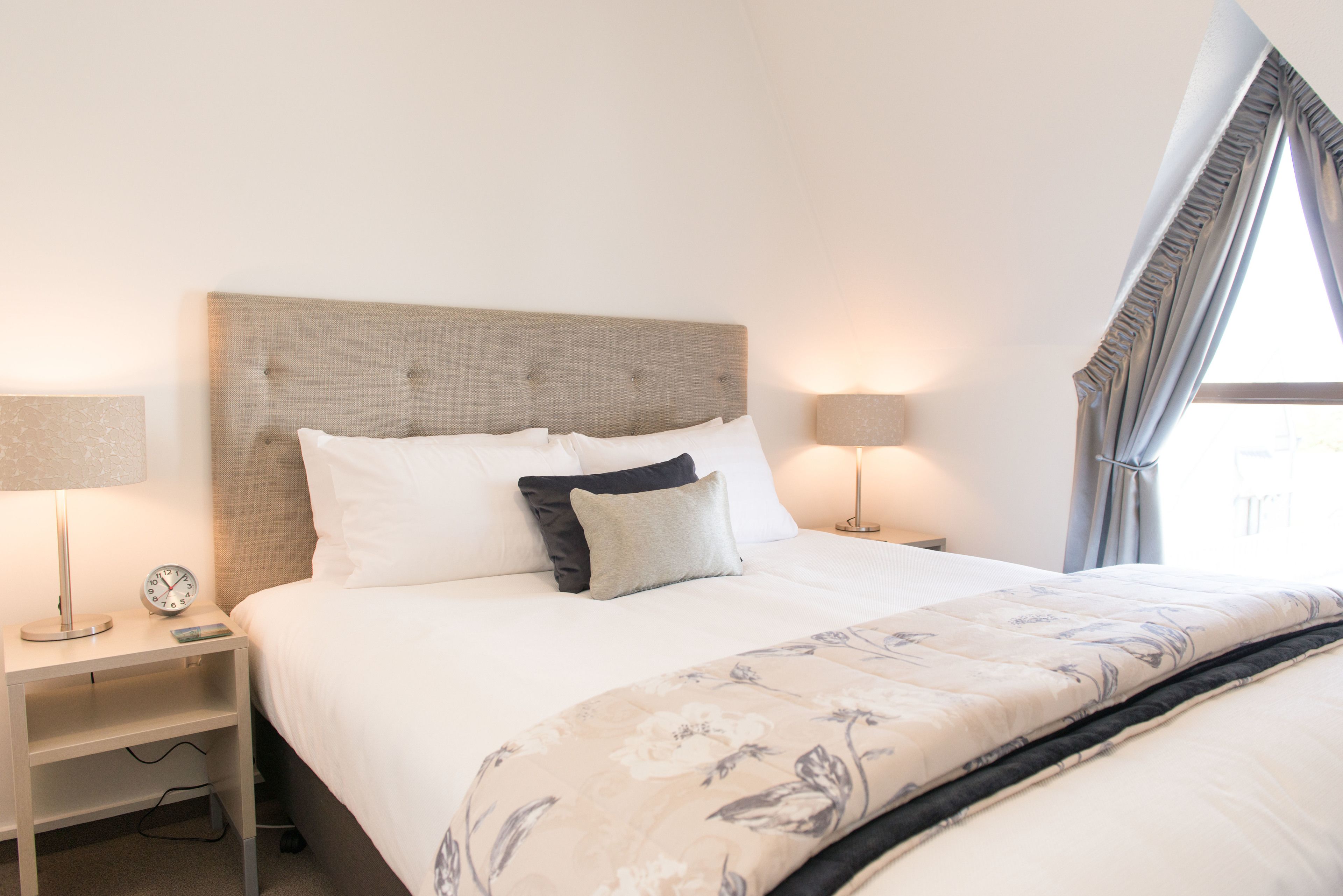 Una cama bien hecha con un edredón floral, dos lámparas de noche, una mesita de noche con un reloj y una ventana con cortinas.