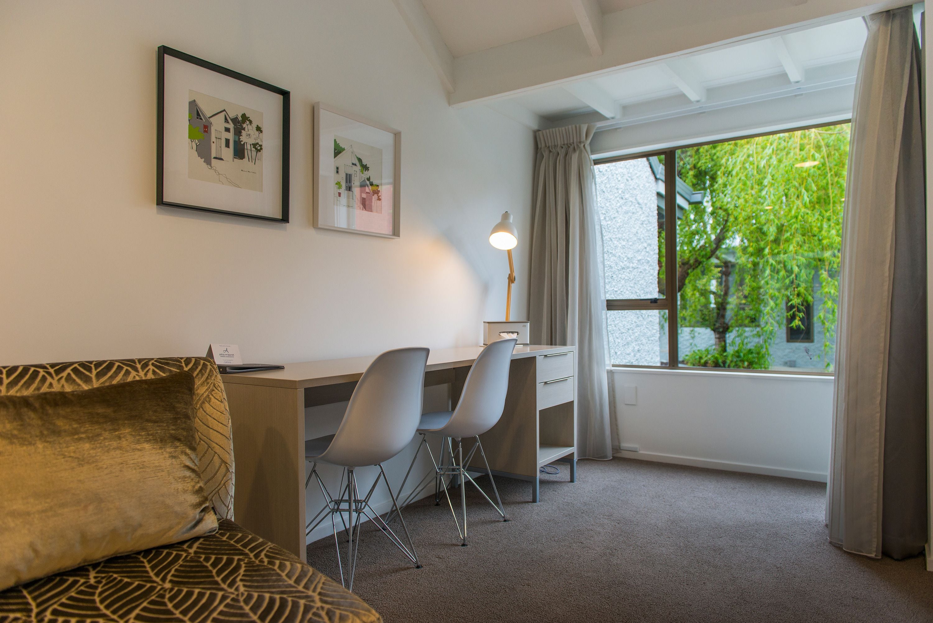 A hotel room with a bed, a desk with two chairs, a lamp, and a window with curtains.