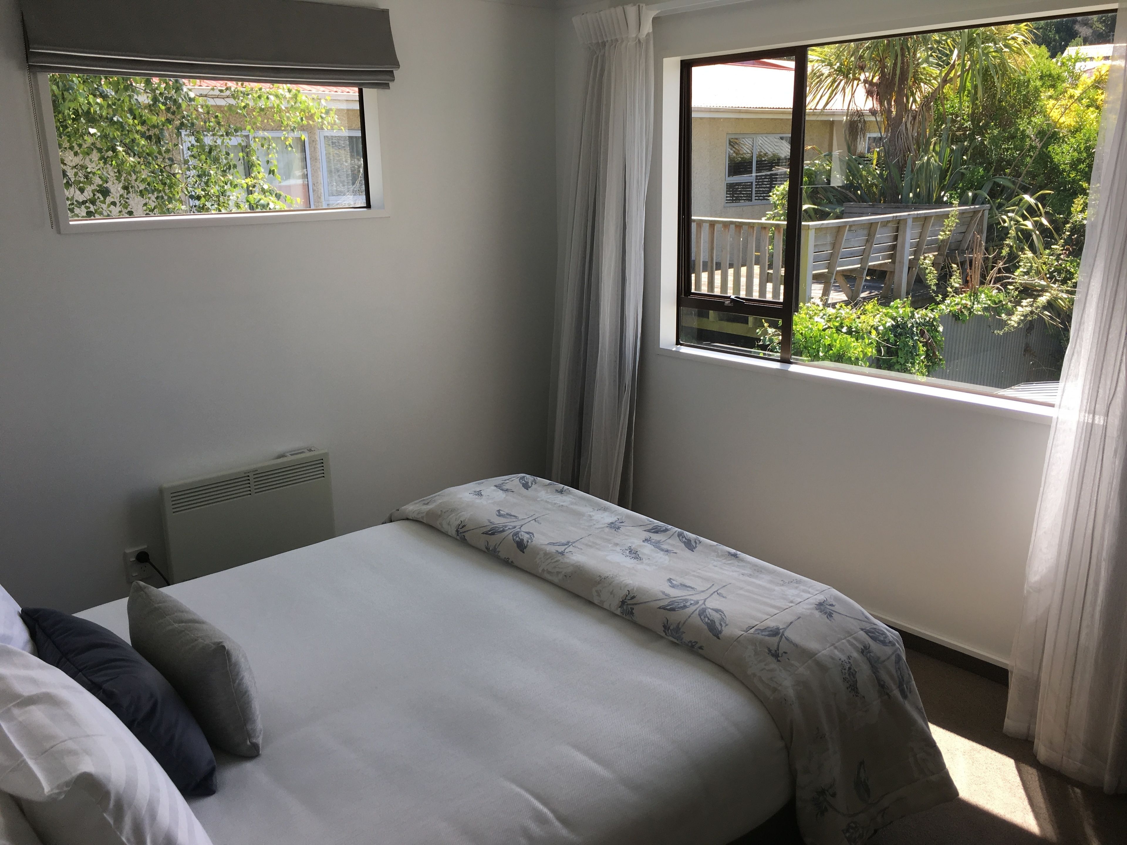 Un dormitorio con cama, un radiador, una ventana con cortinas, y vistas a un balcón y zonas verdes exteriores.
