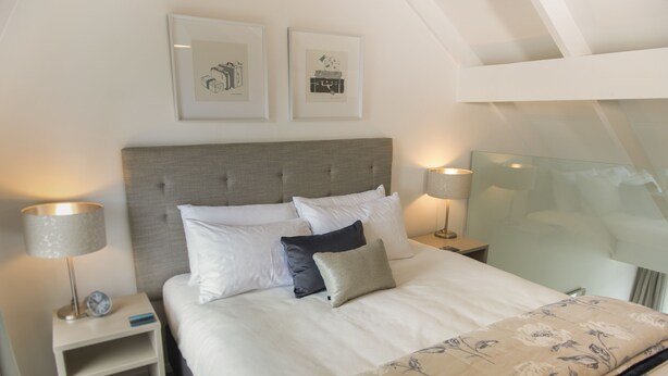 Una cama bien hecha con ropa de cama blanca y gris, dos mesitas de noche con lámparas y cuadros en la pared.