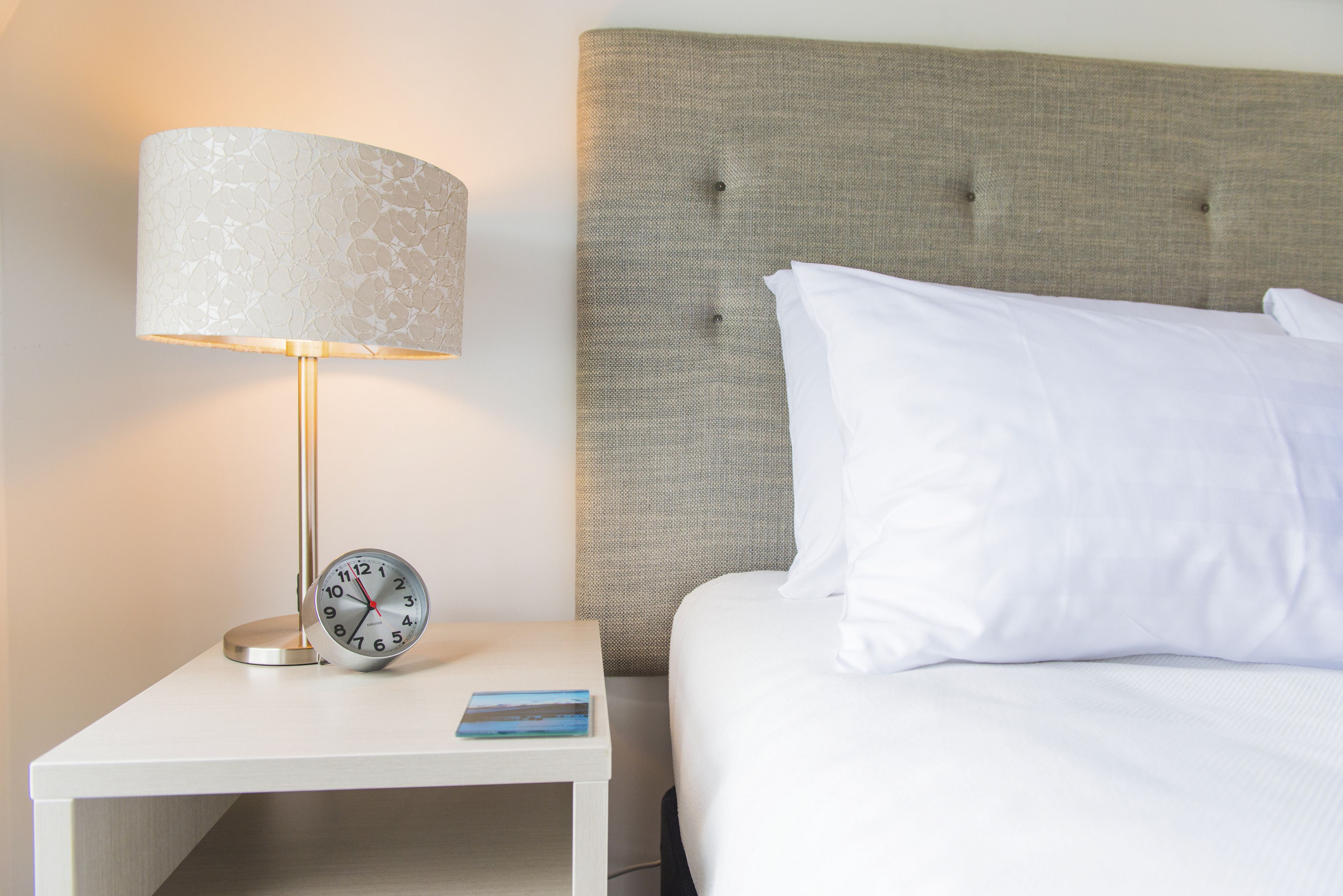 A neatly made bed with white linens, a grey upholstered headboard, a bedside table with a lamp, and a clock.