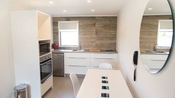 Una cocina moderna con armarios blancos, un horno de acero inoxidable y un lavamanos. Hay una mesa de comedor con sillas y un espejo en la pared.