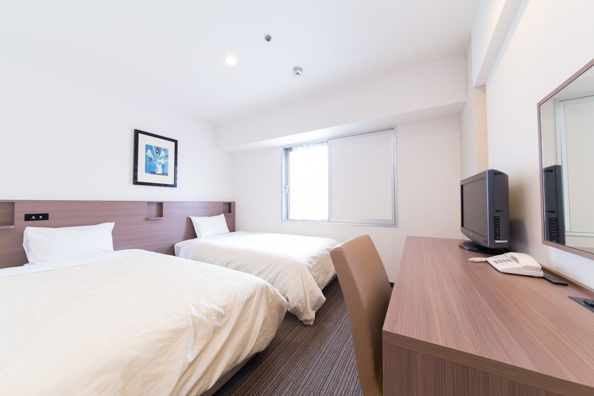 A hotel room with two beds, a desk, a television, and a window.