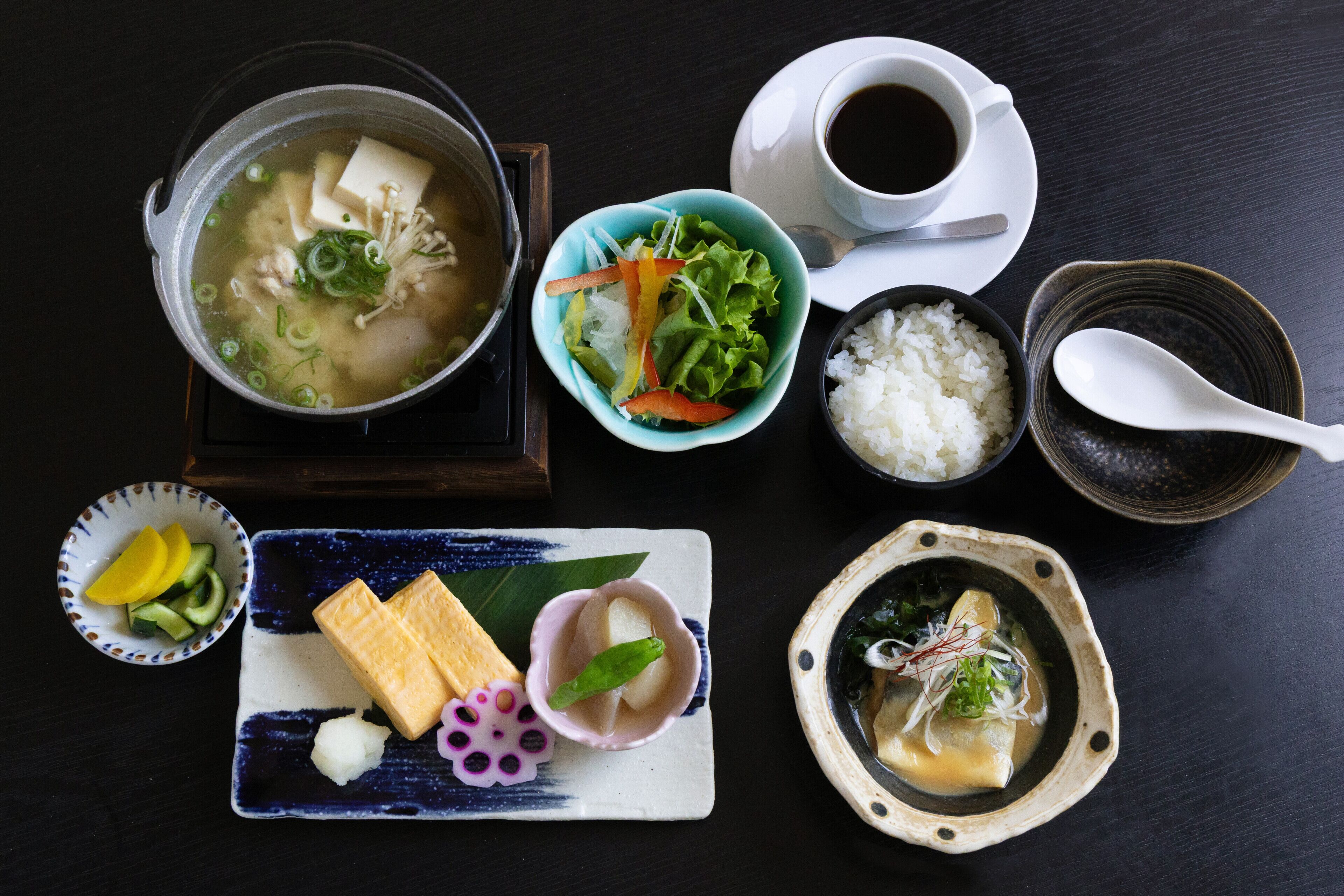 A table with various dishes including a pot of soup, a bowl of rice, a cup of coffee, and several small plates with different foods.