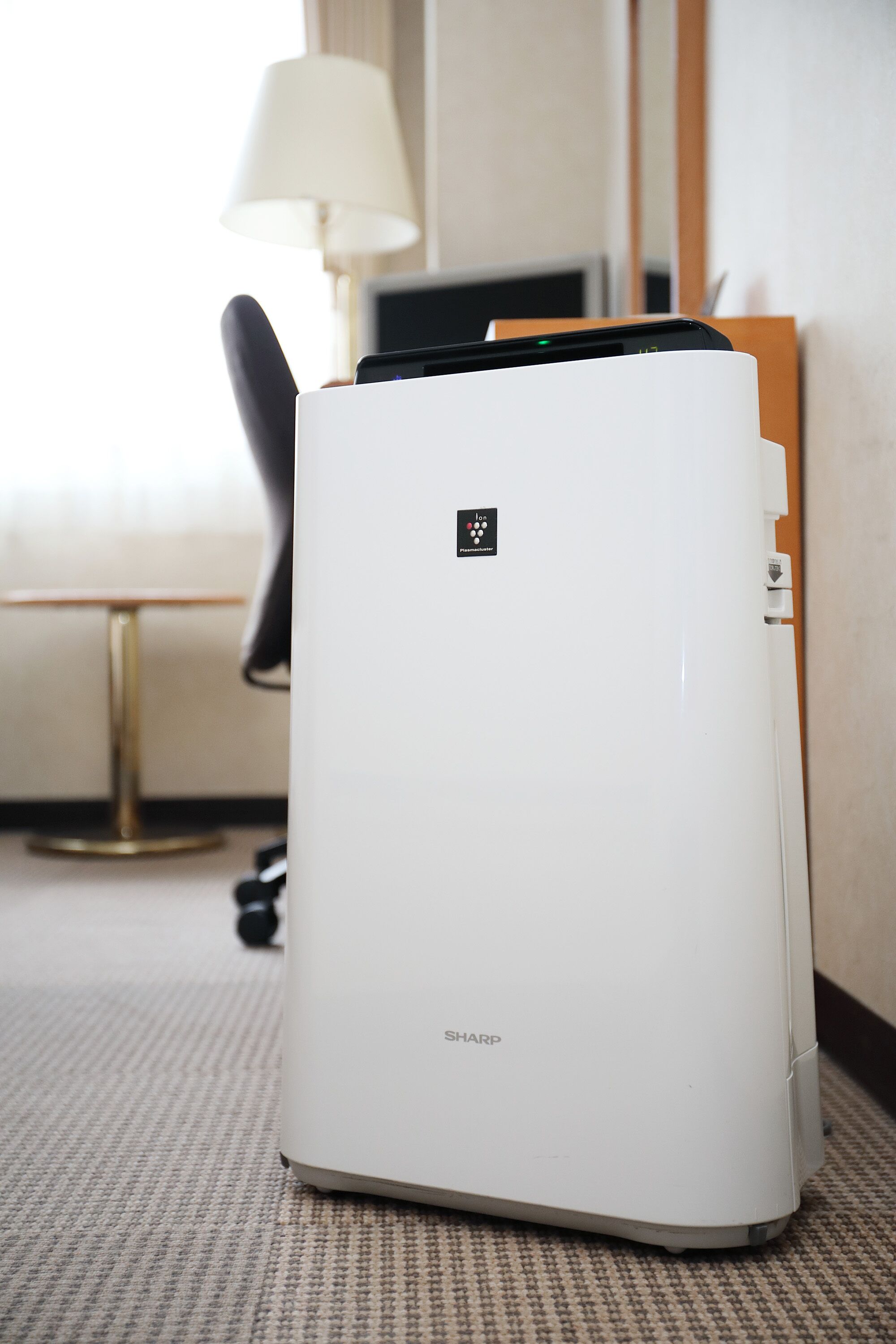 A white Sharp air purifier in a hotel room with a lamp and a chair in the background.