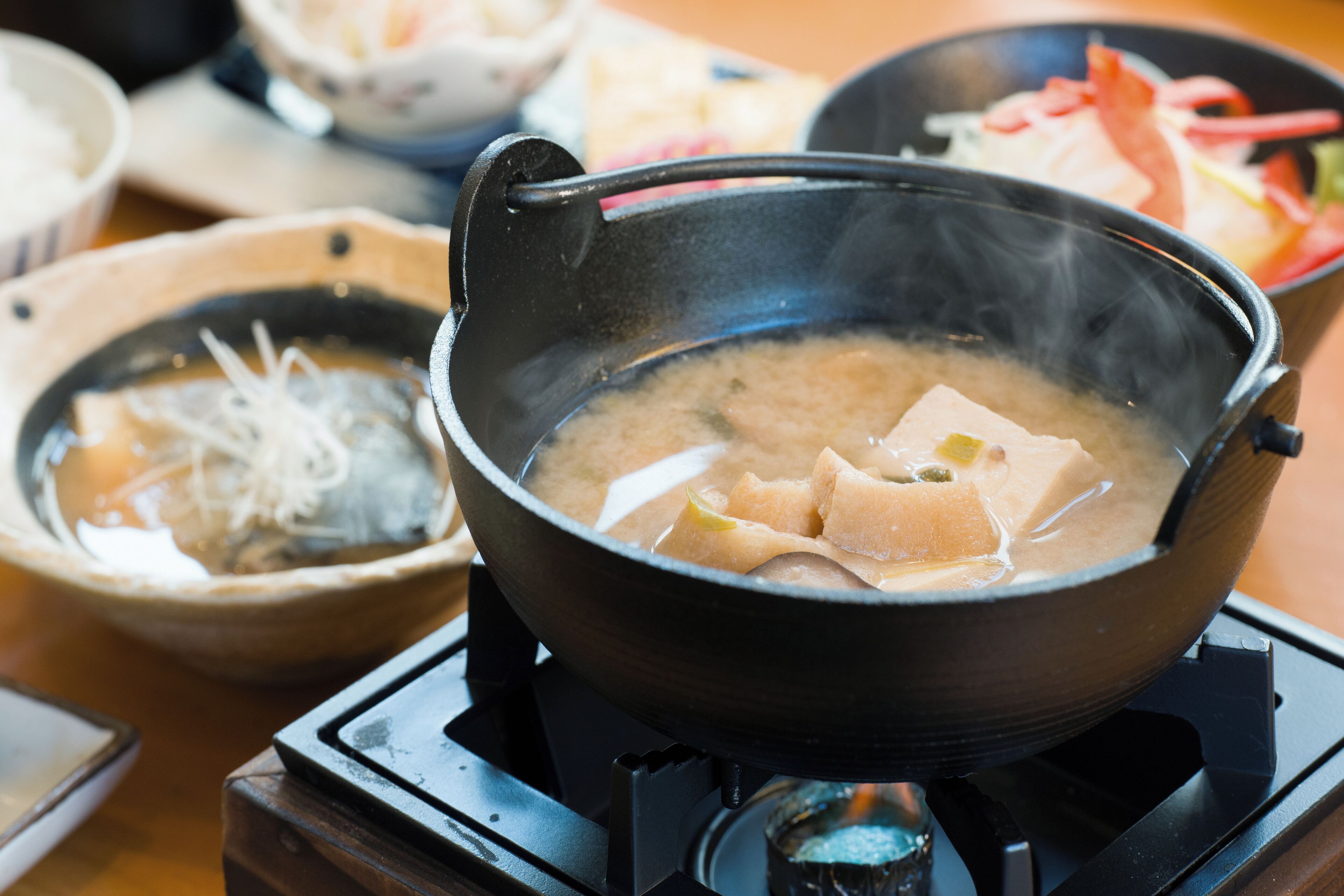 Una olla humeante con tofu y verduras, un tazón de fideos y un plato de carne en rodajas.